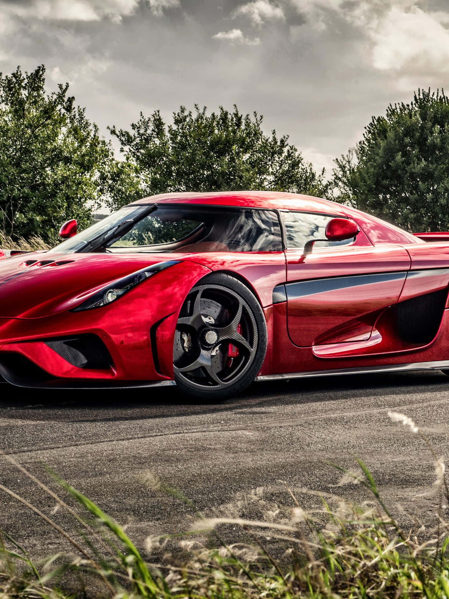 A red Koenigsegg sports car parked on a paved road next to some green grass. The background is blurry and includes trees and a cloudy sky