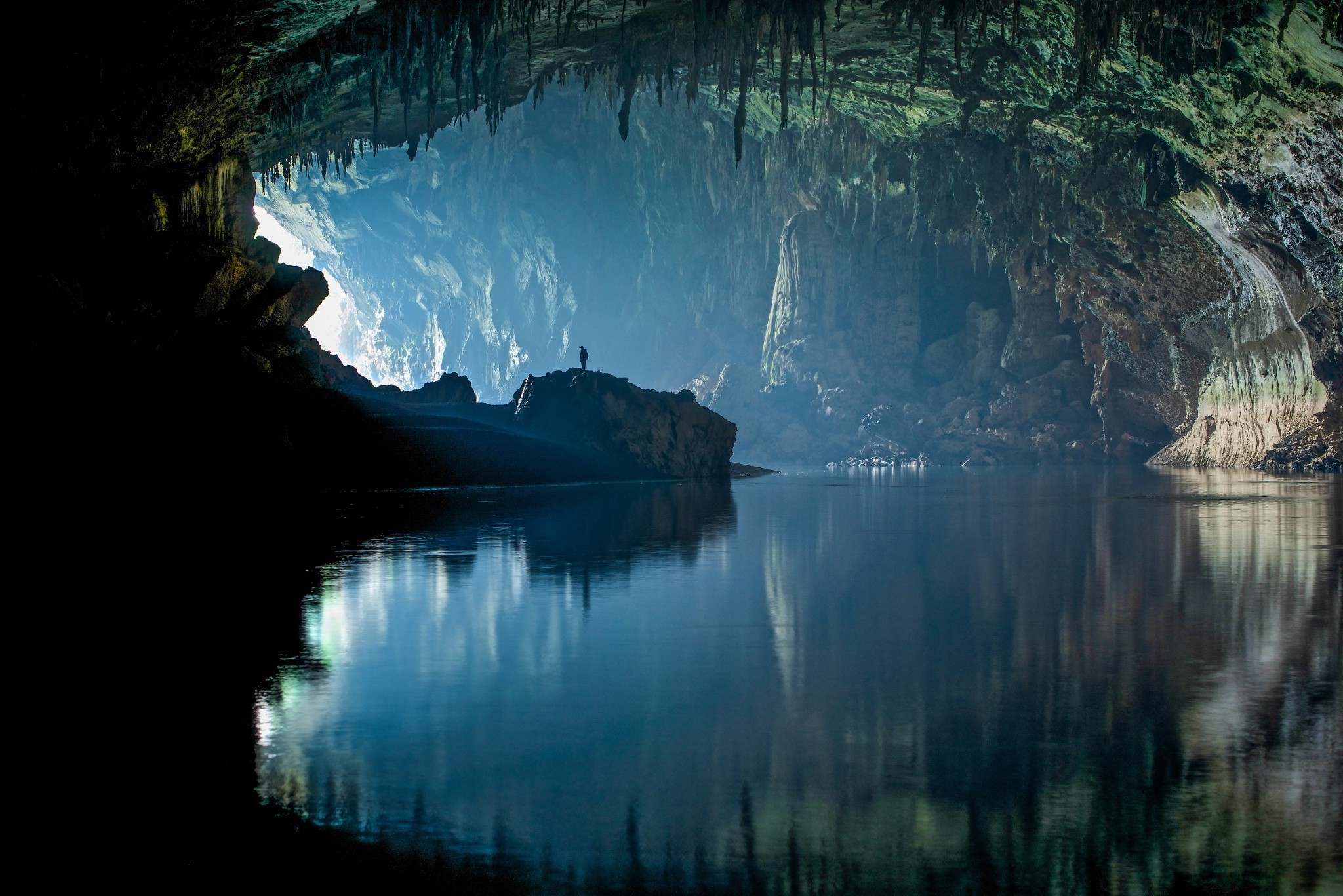 A breathtaking view inside a vast cave, with a person silhouetted against the ethereal light filtering in, a serene underground river reflecting the cave's majestic formations