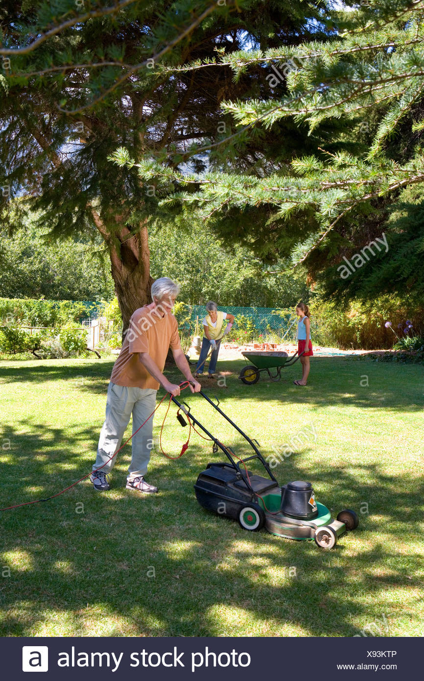 Senior Man Mowing Lawn Wife And Granddaughter With Wheelbarrow In