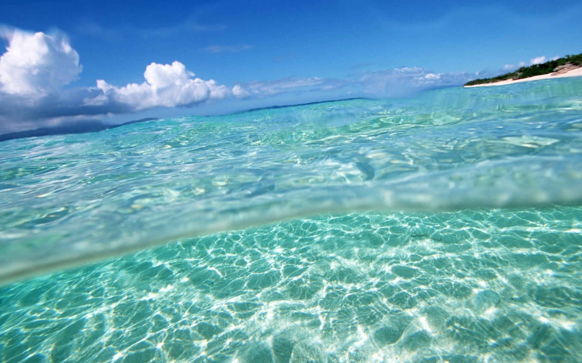 Crystal clear turquoise ocean water gently lapping against a white sand beach under a bright blue sky dotted with fluffy white clouds