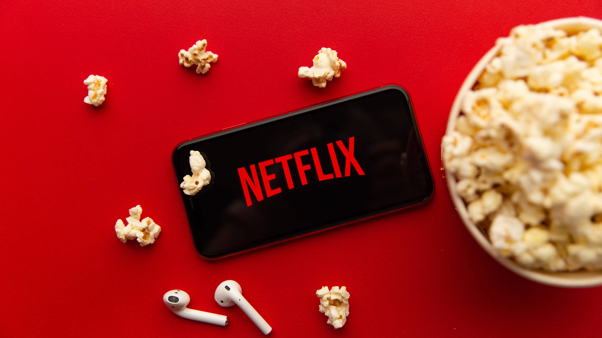A flat lay of a smartphone displaying the Netflix logo, surrounded by popcorn, earbuds, and a bucket of popcorn on a vibrant red background, perfect for Netflix enthusiasts