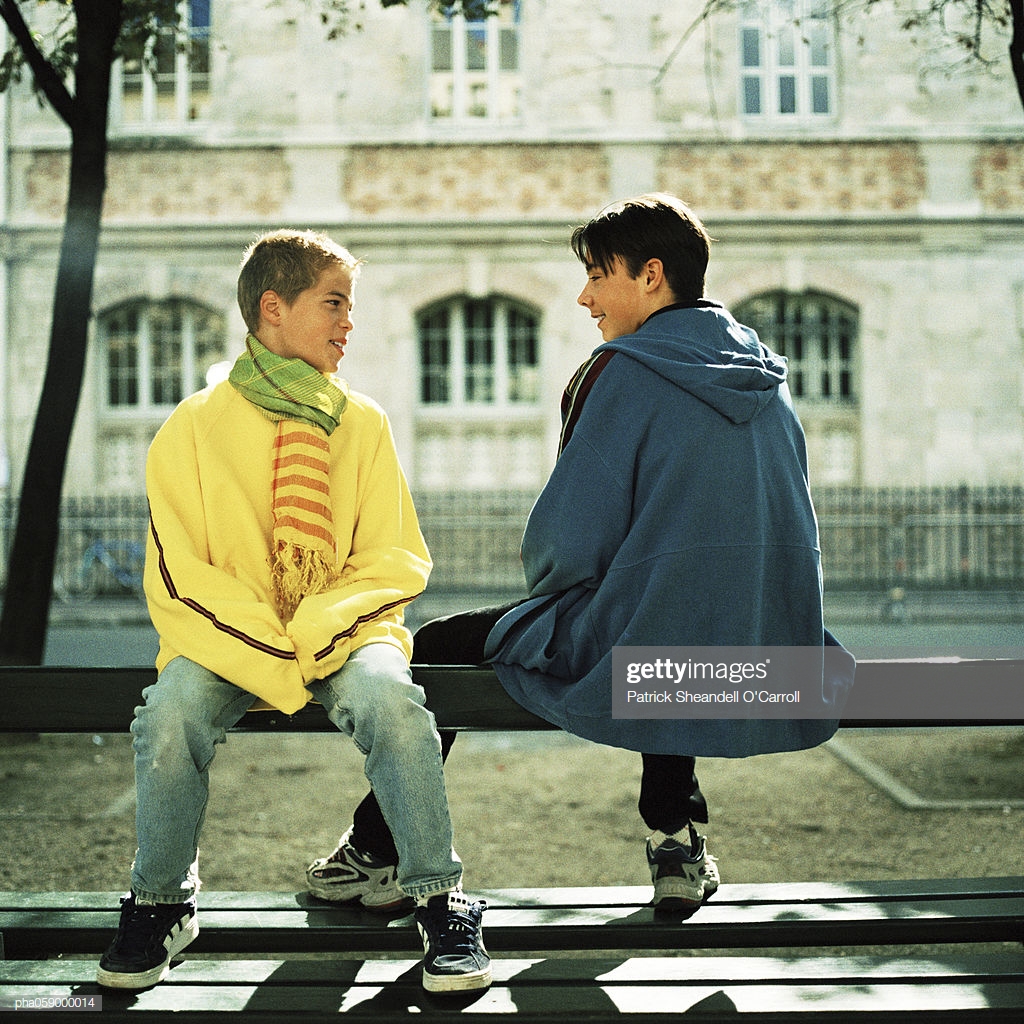 Two Teenage Boys Sitting On Bench Smiling Building In Background