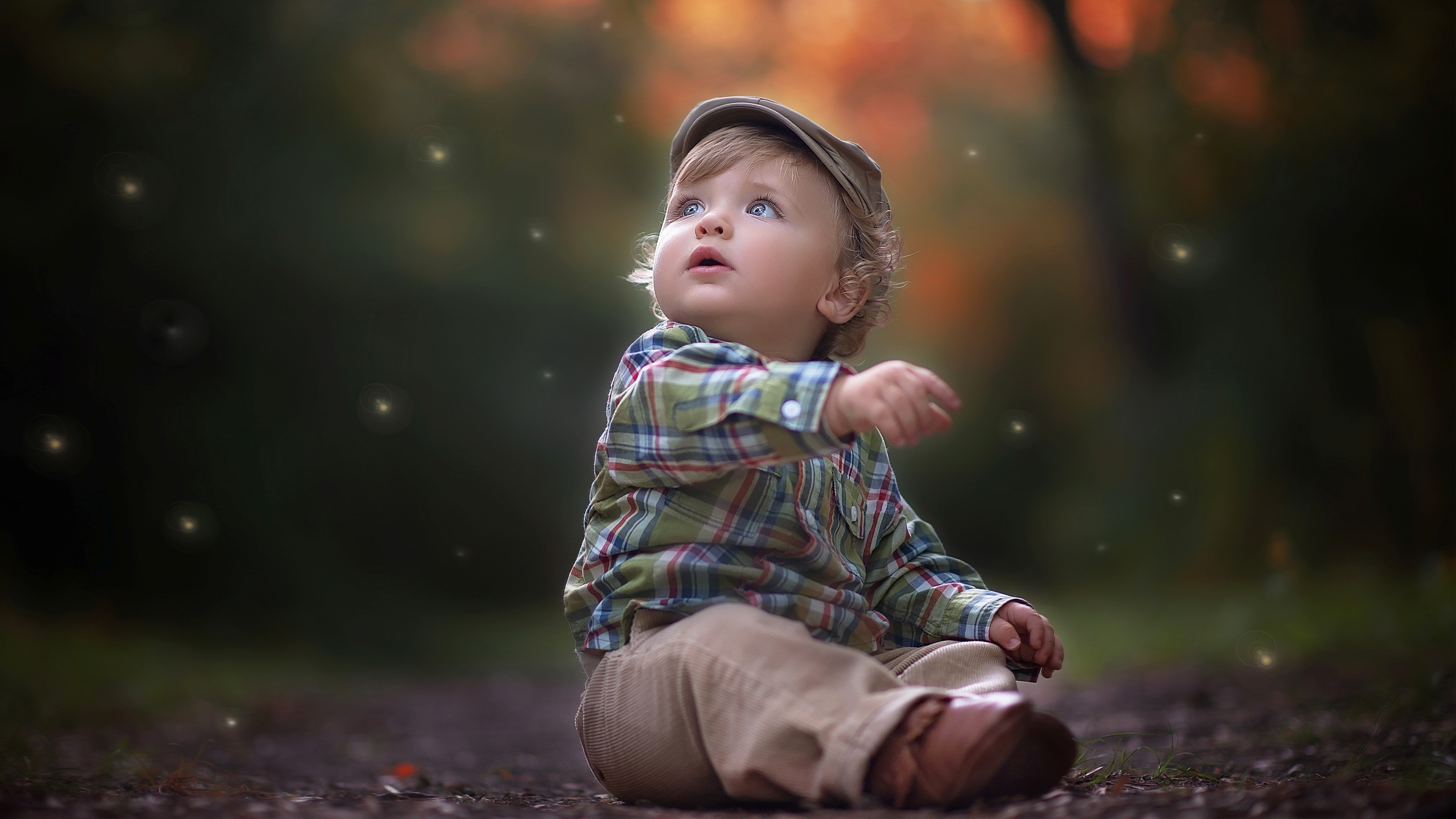 A toddler boy with a cap sits on the ground and looks up at fireflies in a forest