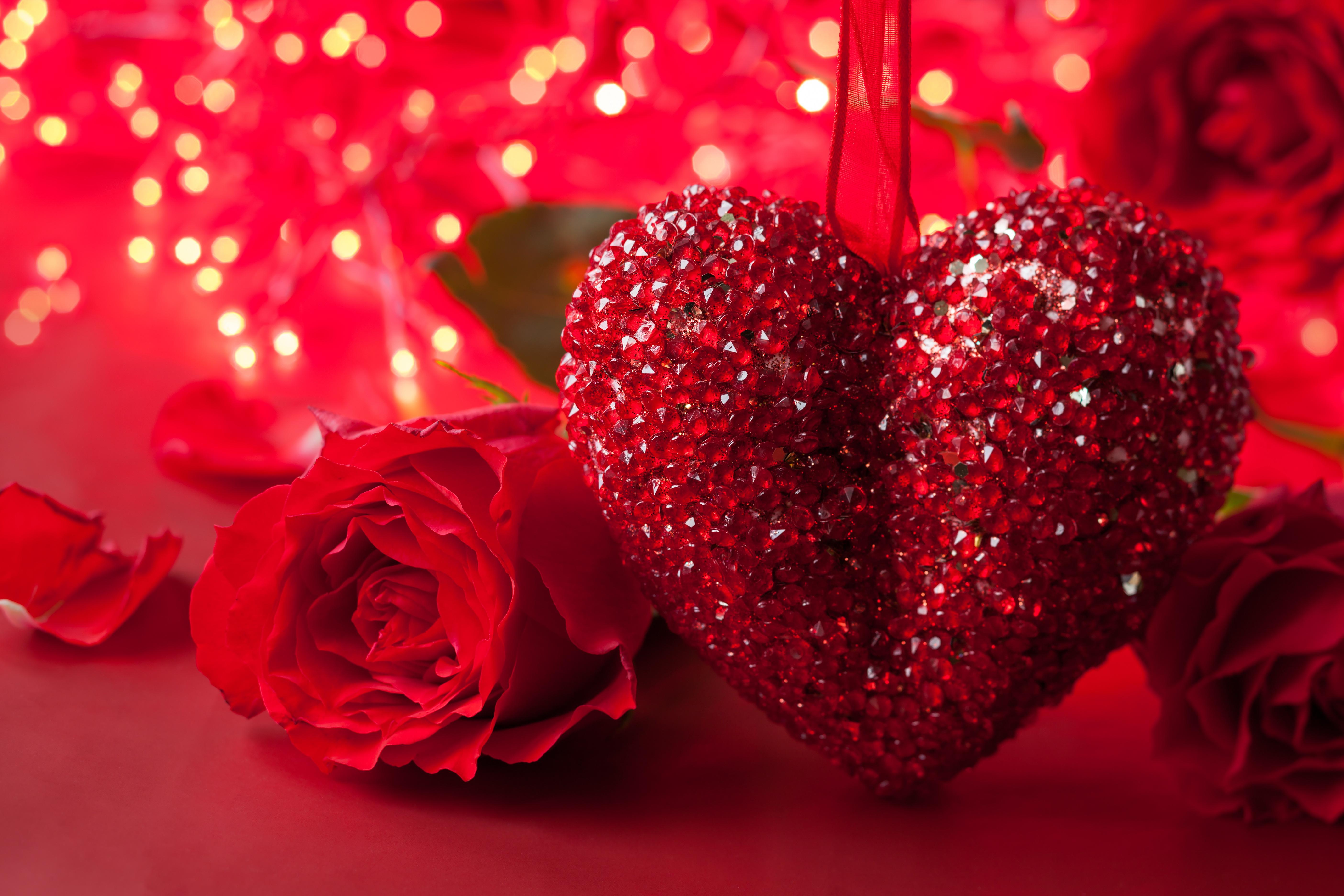 A sparkling red heart ornament hangs amidst red roses, illuminated by a warm, romantic glow of fairy lights, perfect for Valentine's Day or any romantic occasion