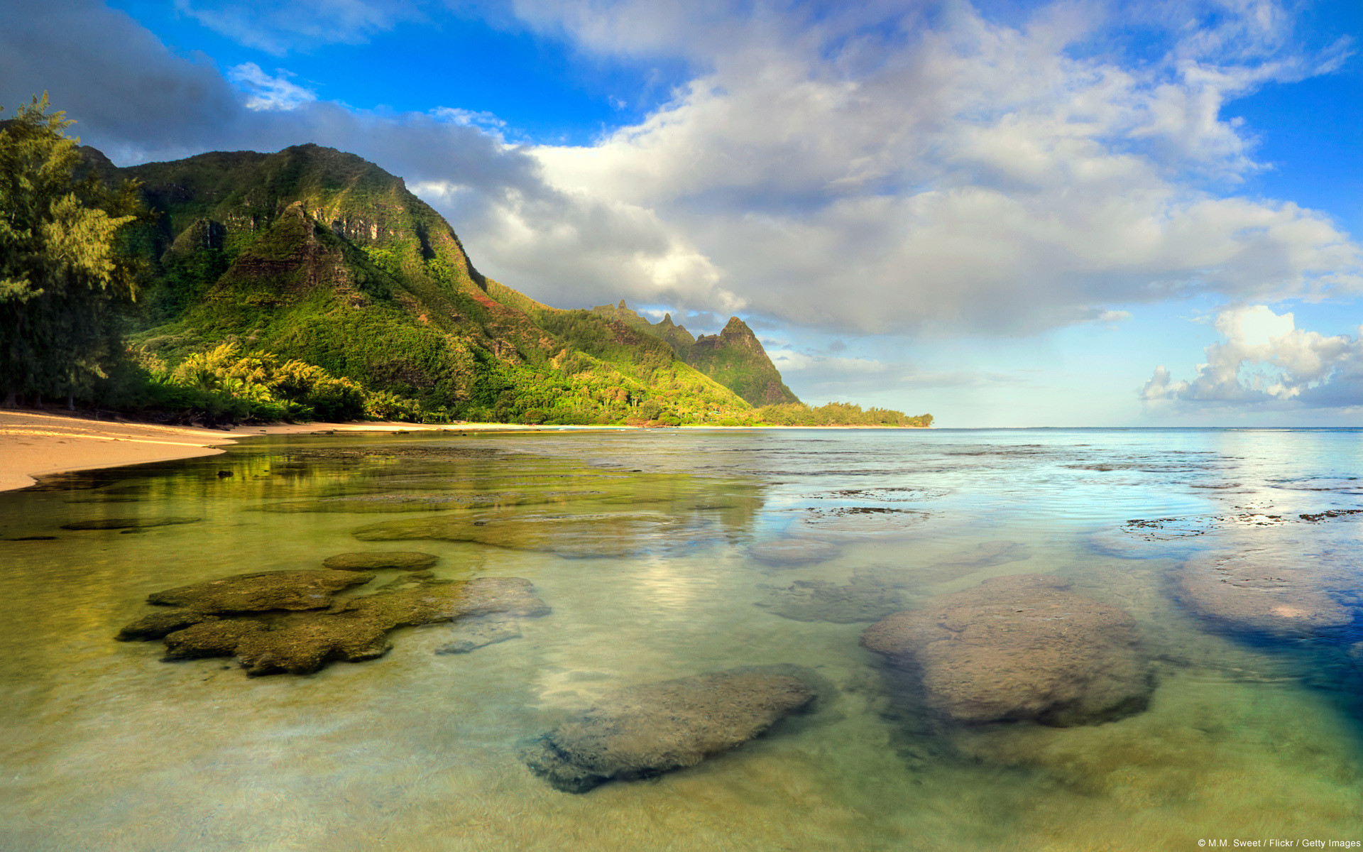 A serene tropical beach with shallow, clear water revealing rocky outcrops, lush green mountains in the background, and a cloudy blue sky