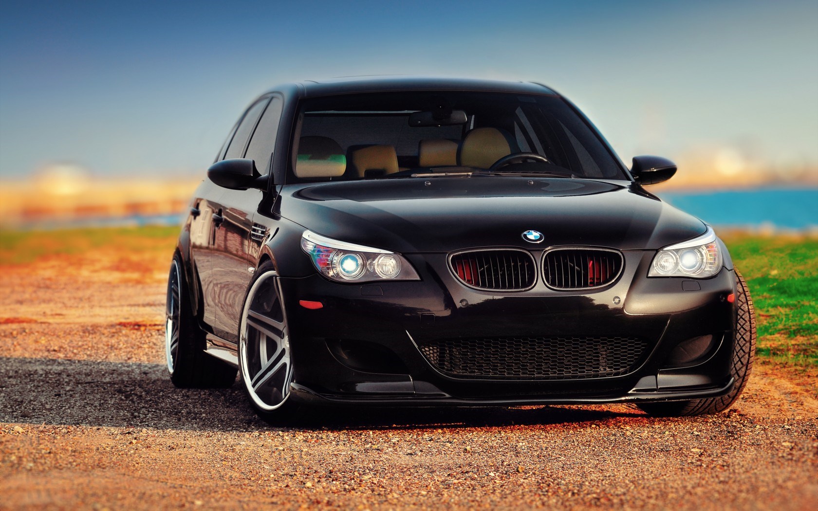 Black BMW M5 parked on a dirt road, overlooking a body of water at sunset