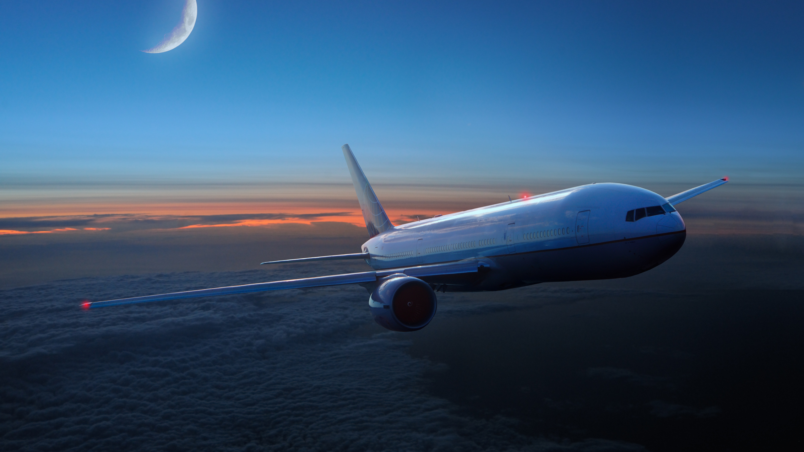 A passenger airliner soaring through the twilight sky above a layer of clouds, with a crescent moon visible in the background