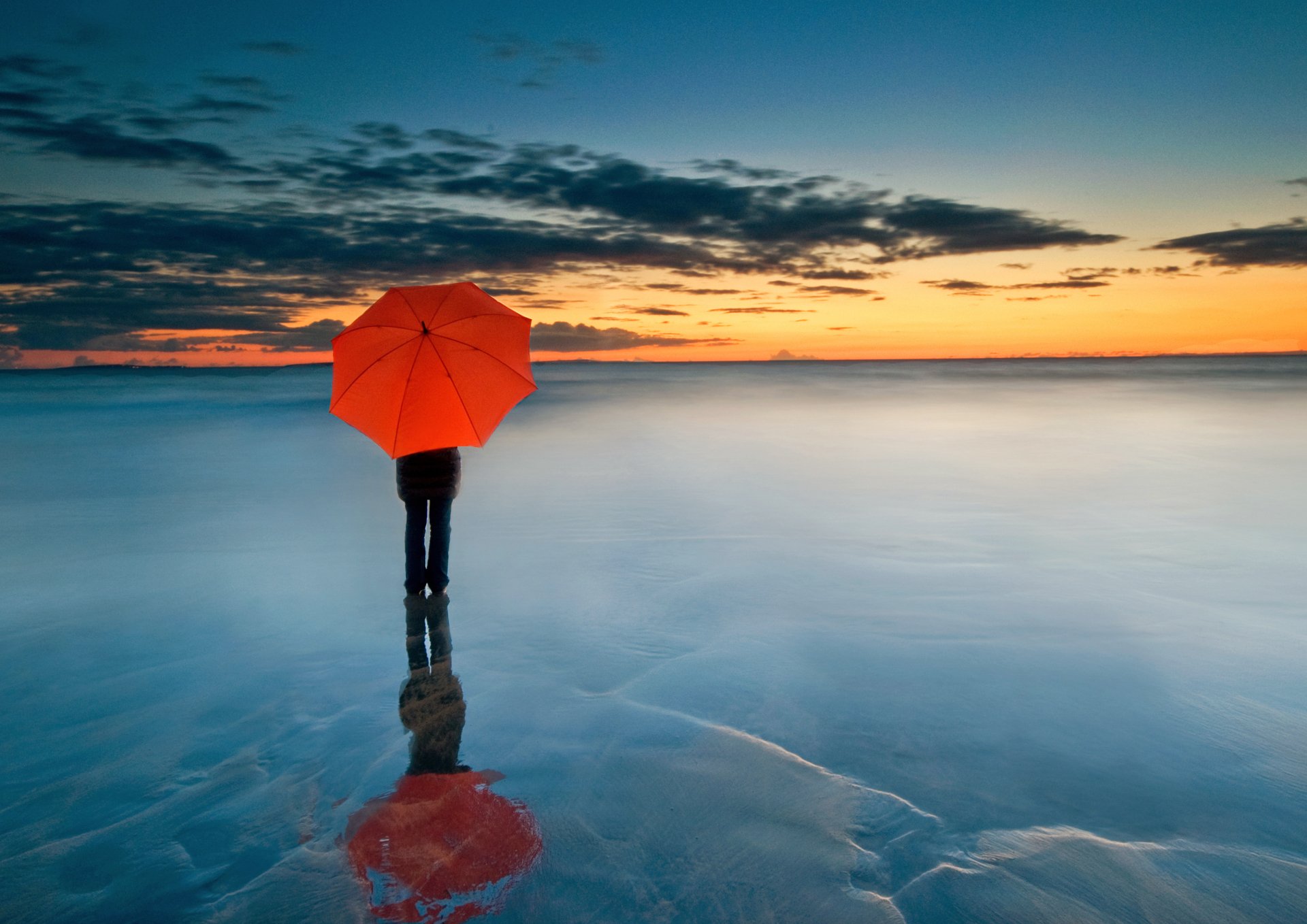 A lone figure with a red umbrella stands on a serene beach at sunset, their reflection mirrored in the wet sand. The smooth water and vibrant sky create a tranquil and picturesque scene