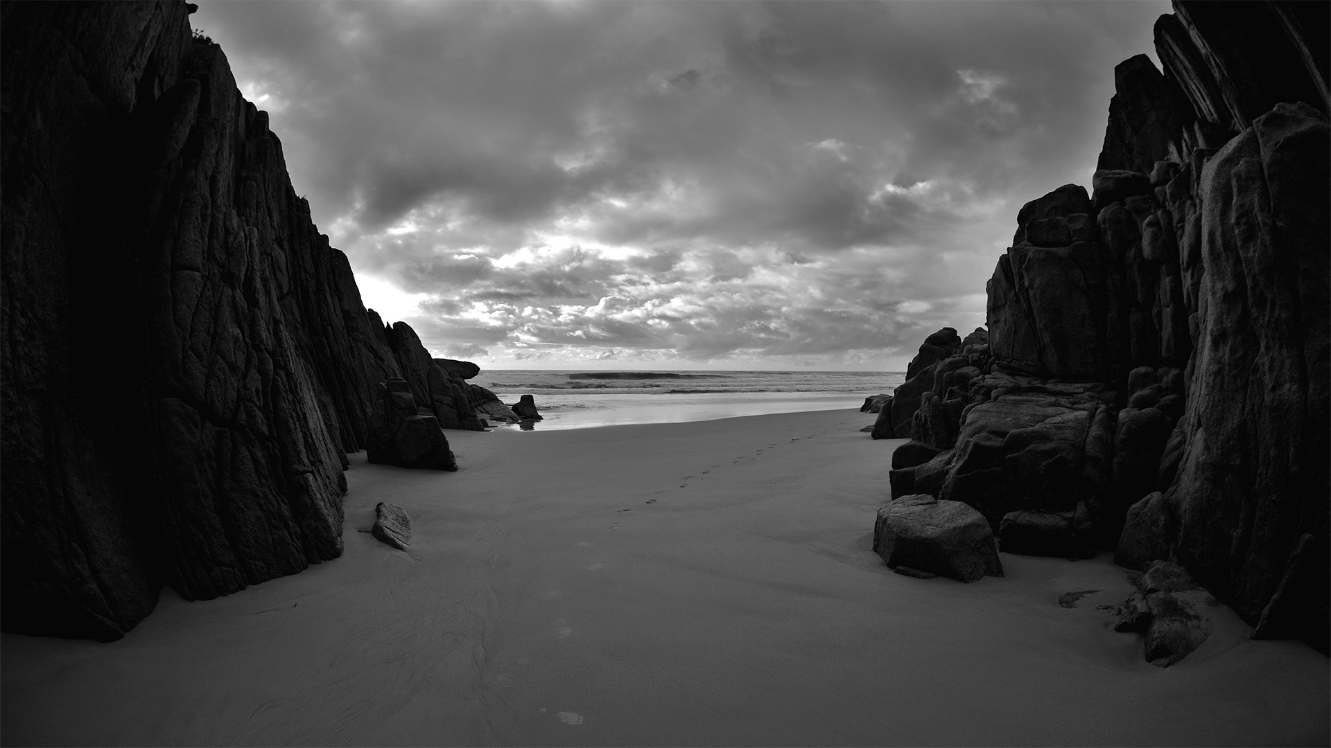 Black and white photograph of a secluded beach, framed by dark, rocky cliffs. Footprints trail across the sand towards the ocean under a cloudy sky