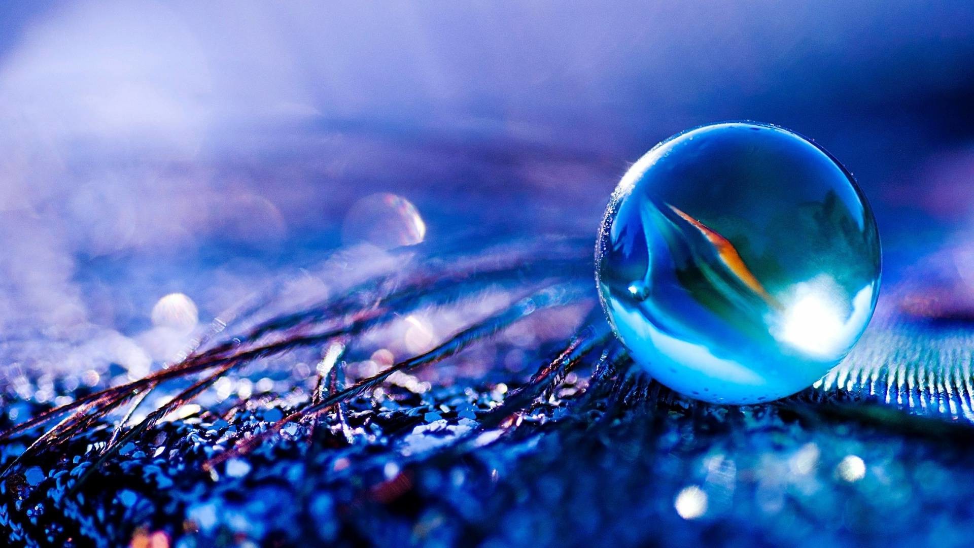 A close-up, macro photograph of a blue marble resting on a textured, glittery surface with feather-like strands. The backdrop is a soft, defocused blue