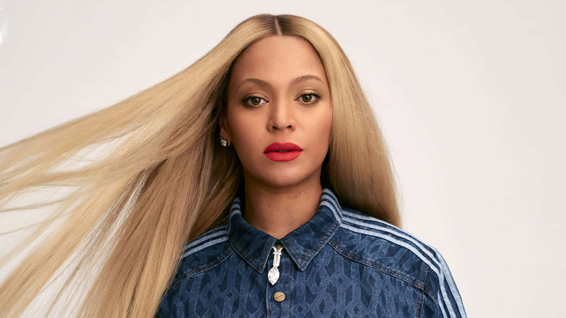 Beyoncé Knowles-Carter with long, flowing blonde hair, wearing a denim shirt with a diamond pendant, against a light background