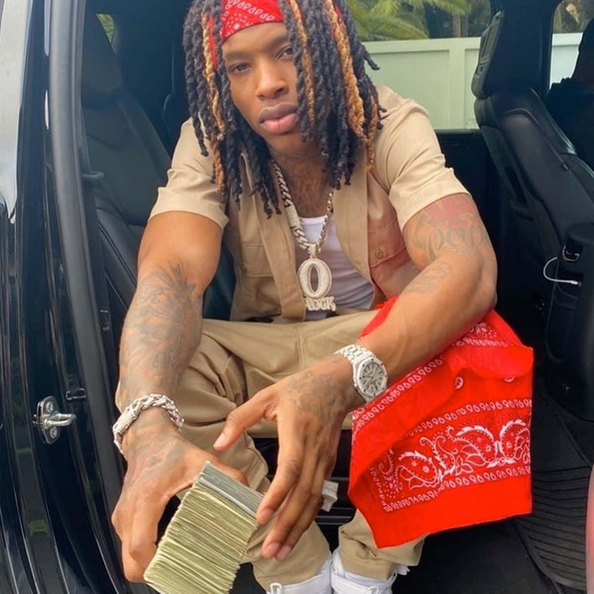 A man sitting in a car, holding a stack of money and a red bandana
