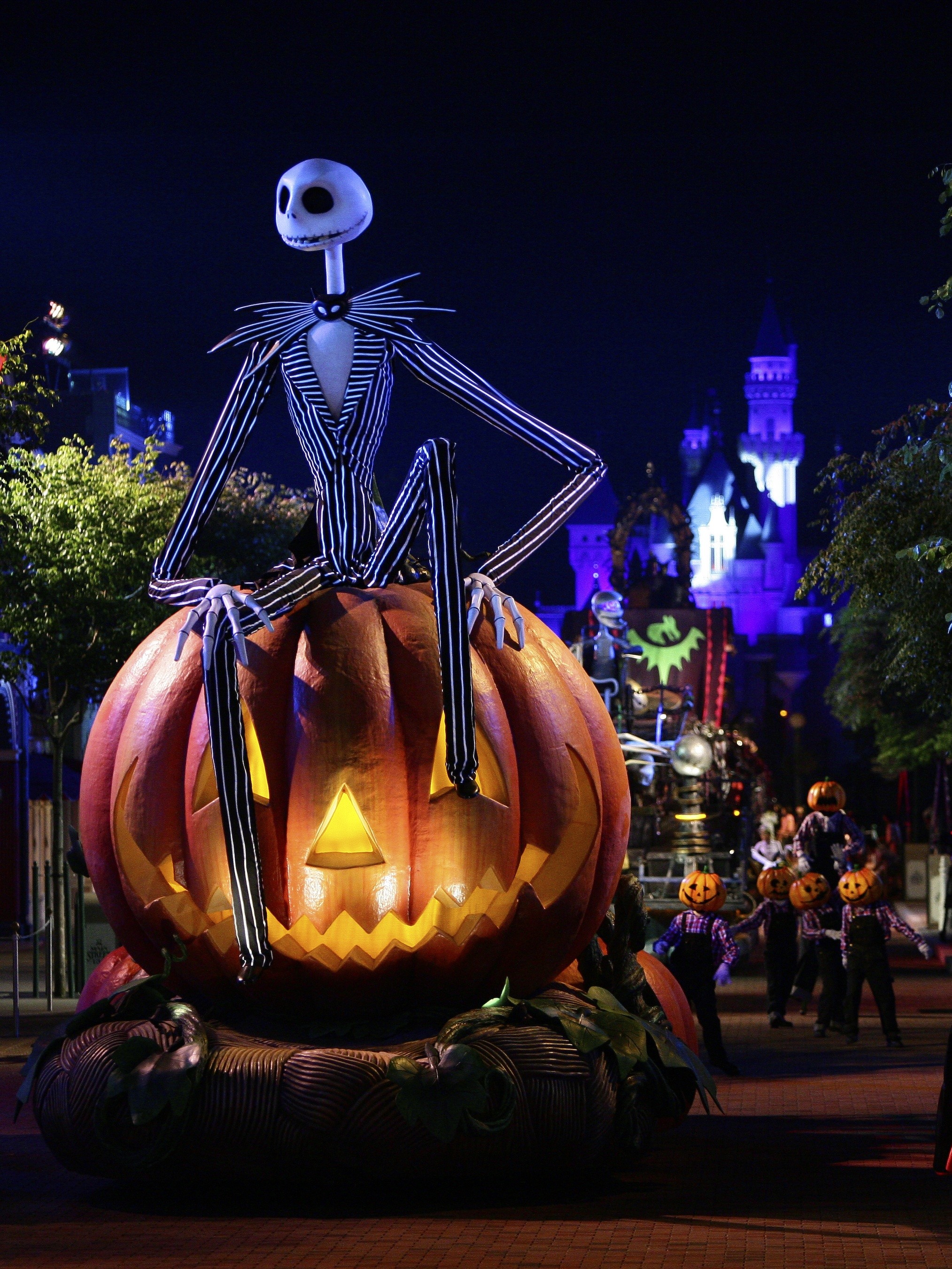 Jack Skellington sitting on a giant, carved pumpkin in front of a Halloween-decorated Disney castle at night