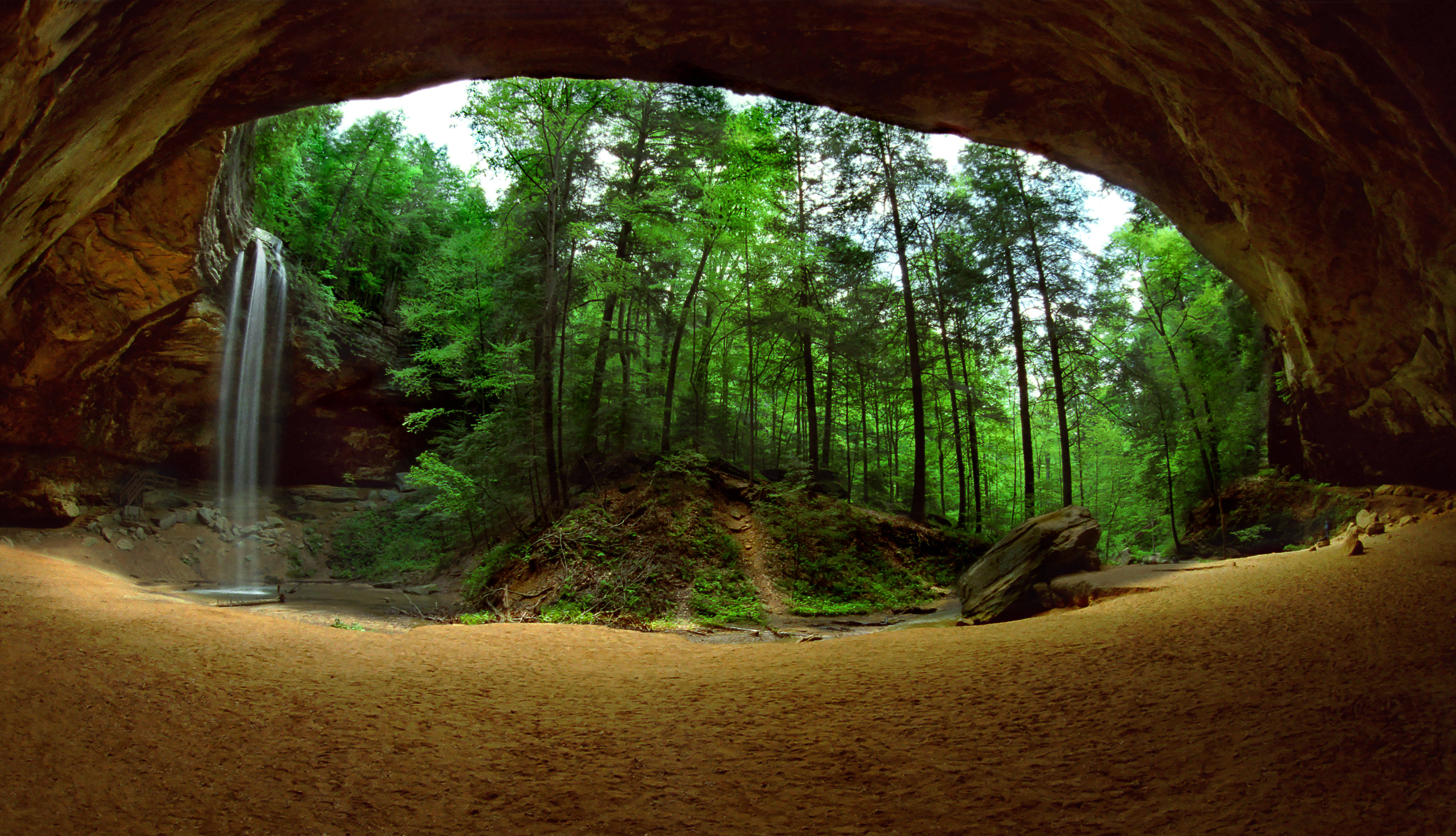 A captivating view from inside a cave, showcasing a waterfall cascading into a sandy area, surrounded by lush green trees and foliage, creating a serene nature background