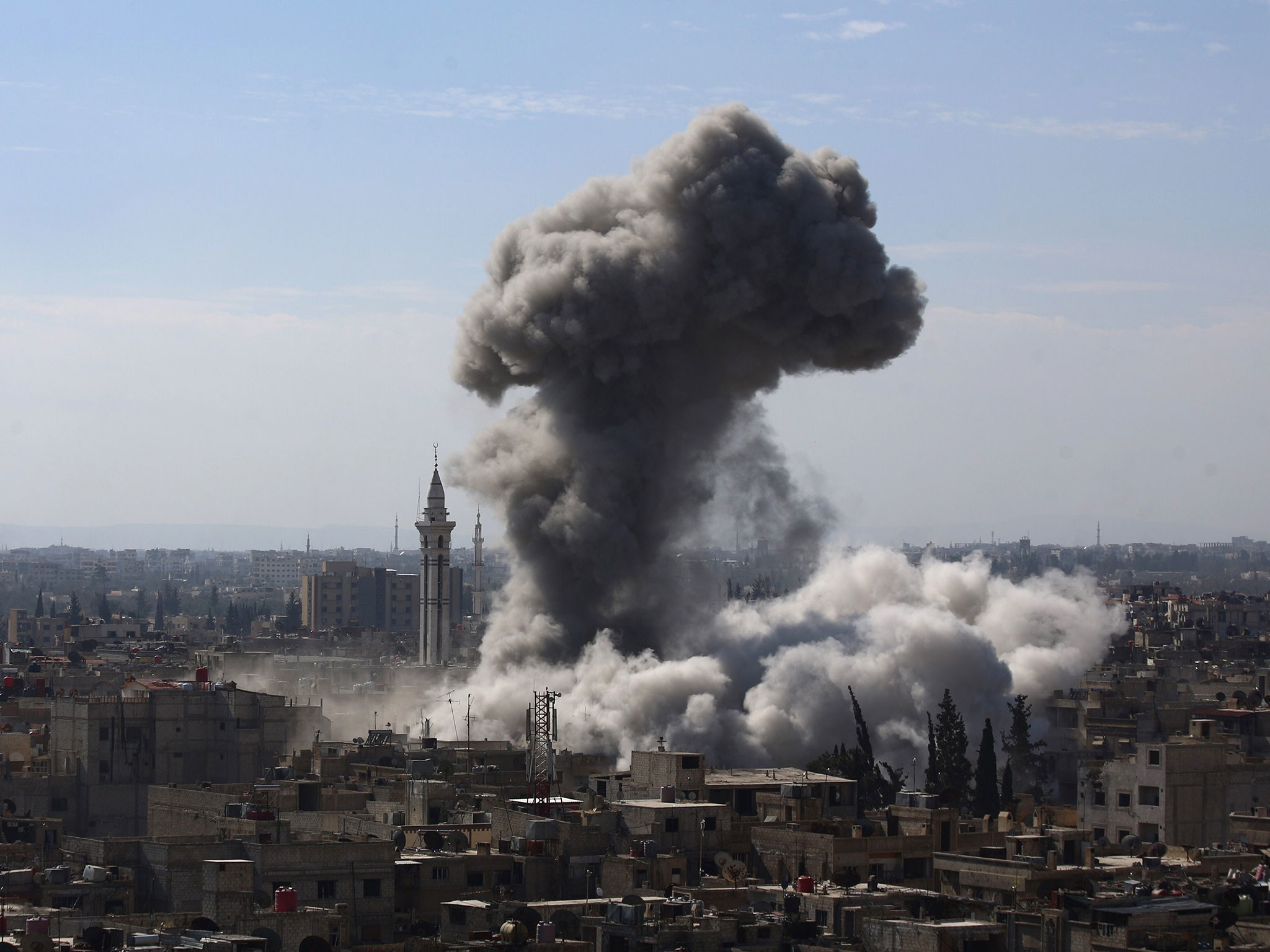 A massive cloud of dark smoke billows over a densely populated Syrian city, indicating a recent explosion or airstrike. Buildings and a mosque are visible amidst the destruction