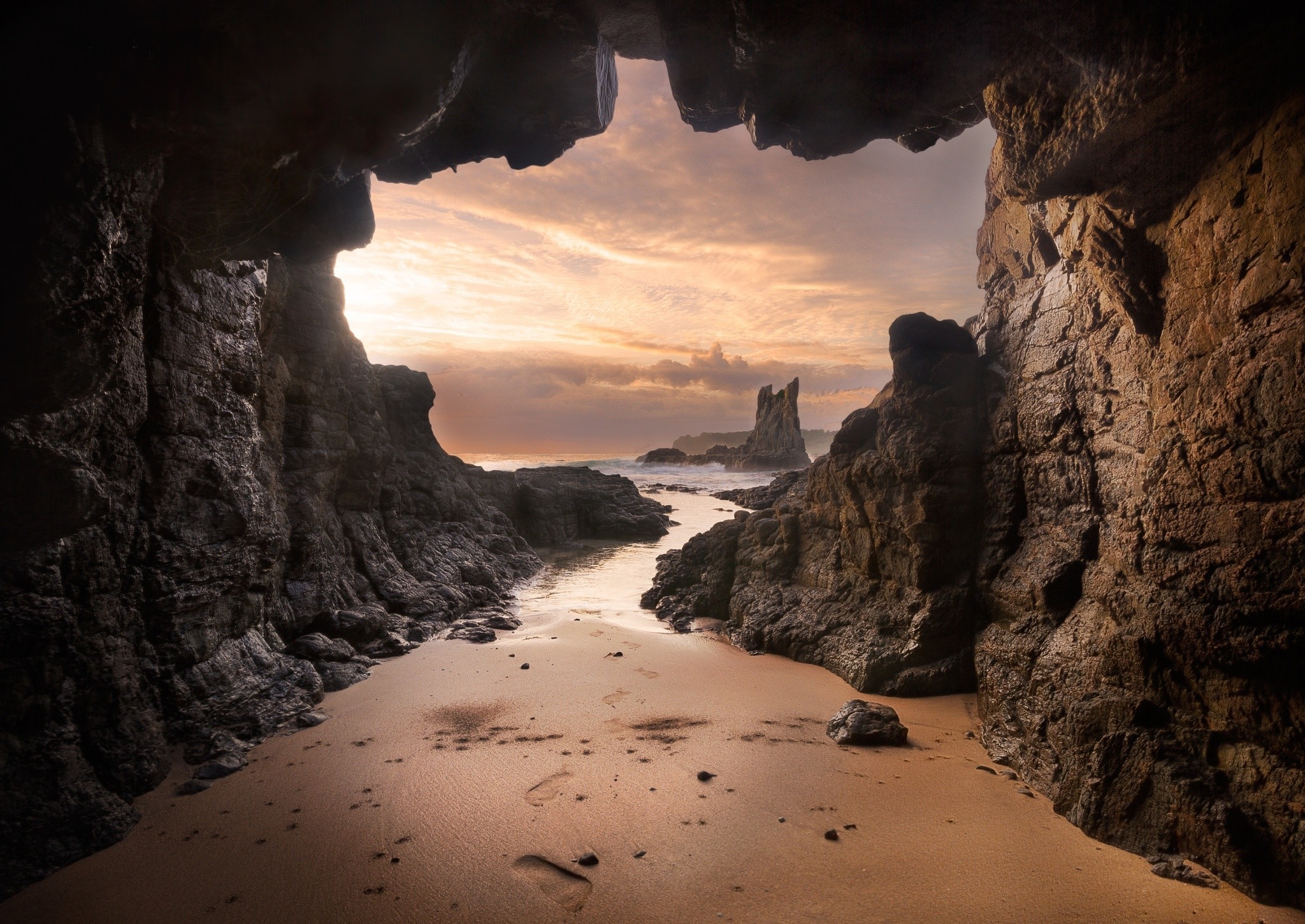 A breathtaking view of the sunrise through a sea cave, with the sandy beach in the foreground and rocky formations leading to the ocean