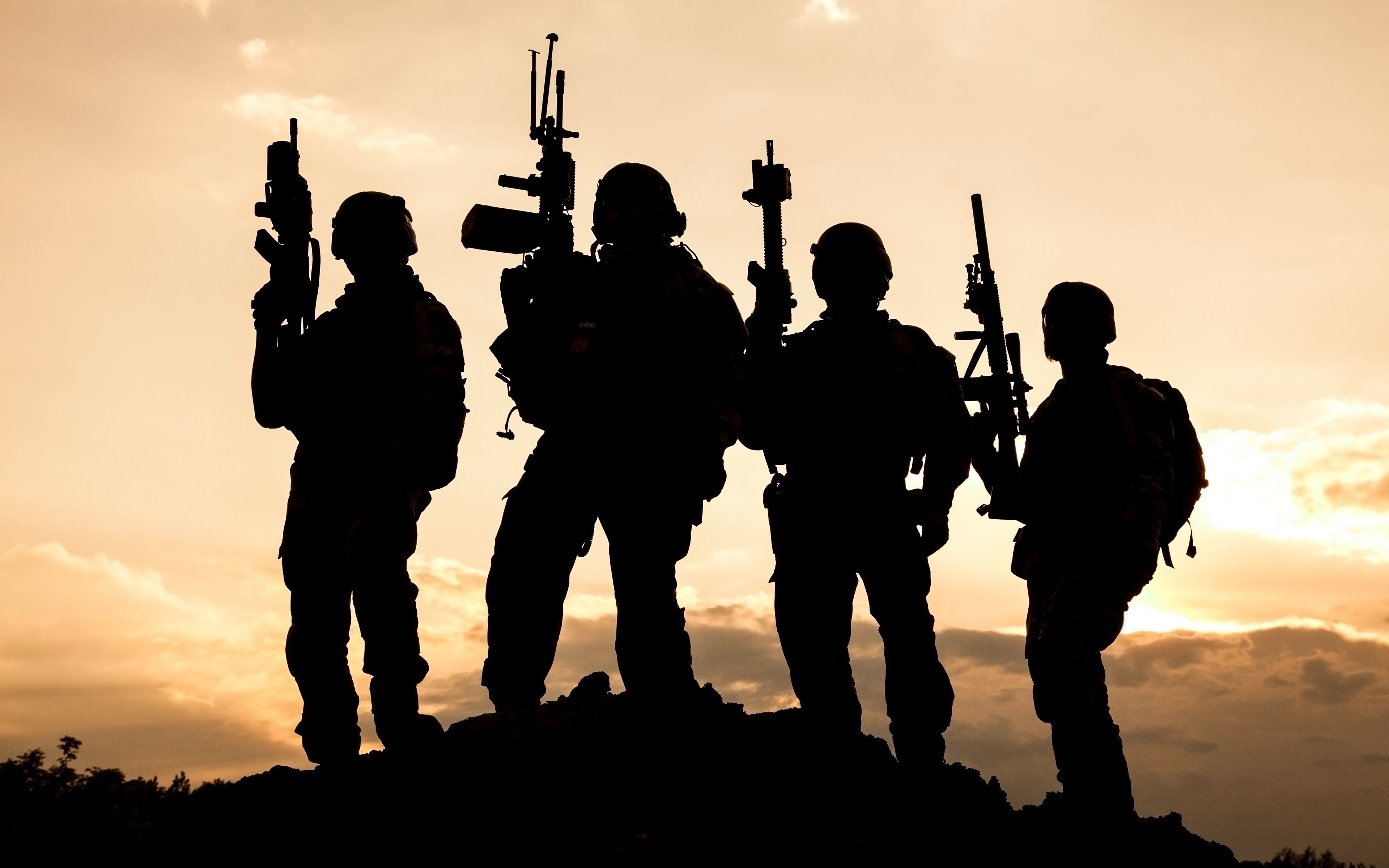 Silhouettes of four soldiers standing on a hilltop against a sunset backdrop, holding various firearms