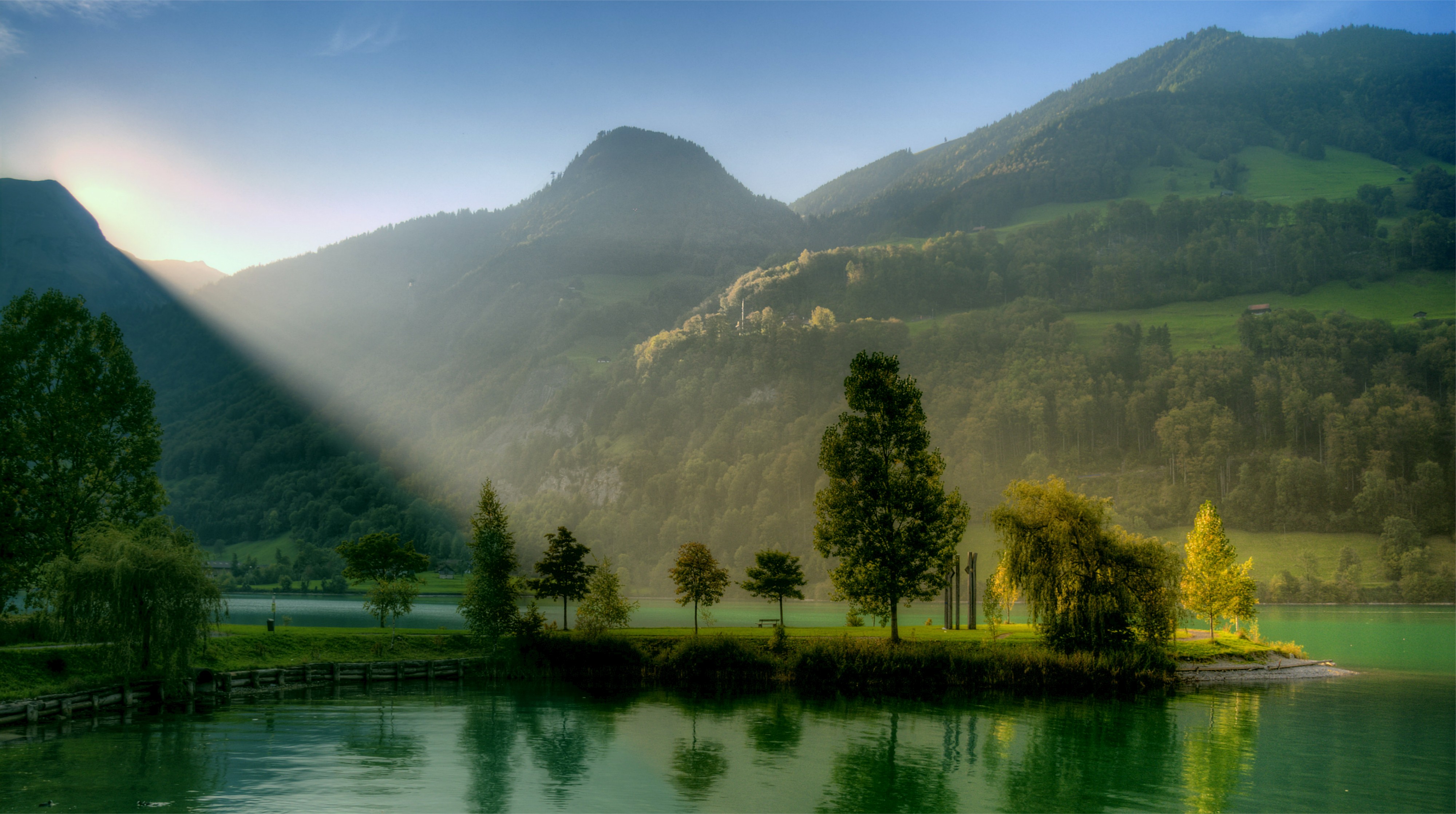 Serene landscape with a lake reflecting sunlight, surrounded by lush green trees and rolling hills
