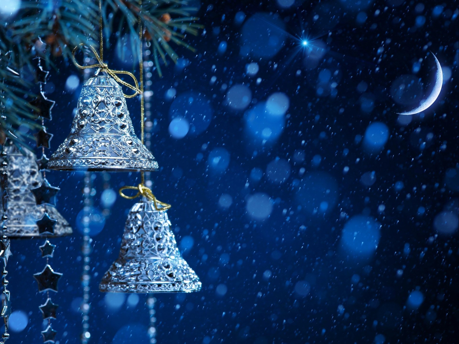 Silver Christmas bells hanging on a snow-dusted fir tree branch against a dark blue night sky with a crescent moon and falling snow