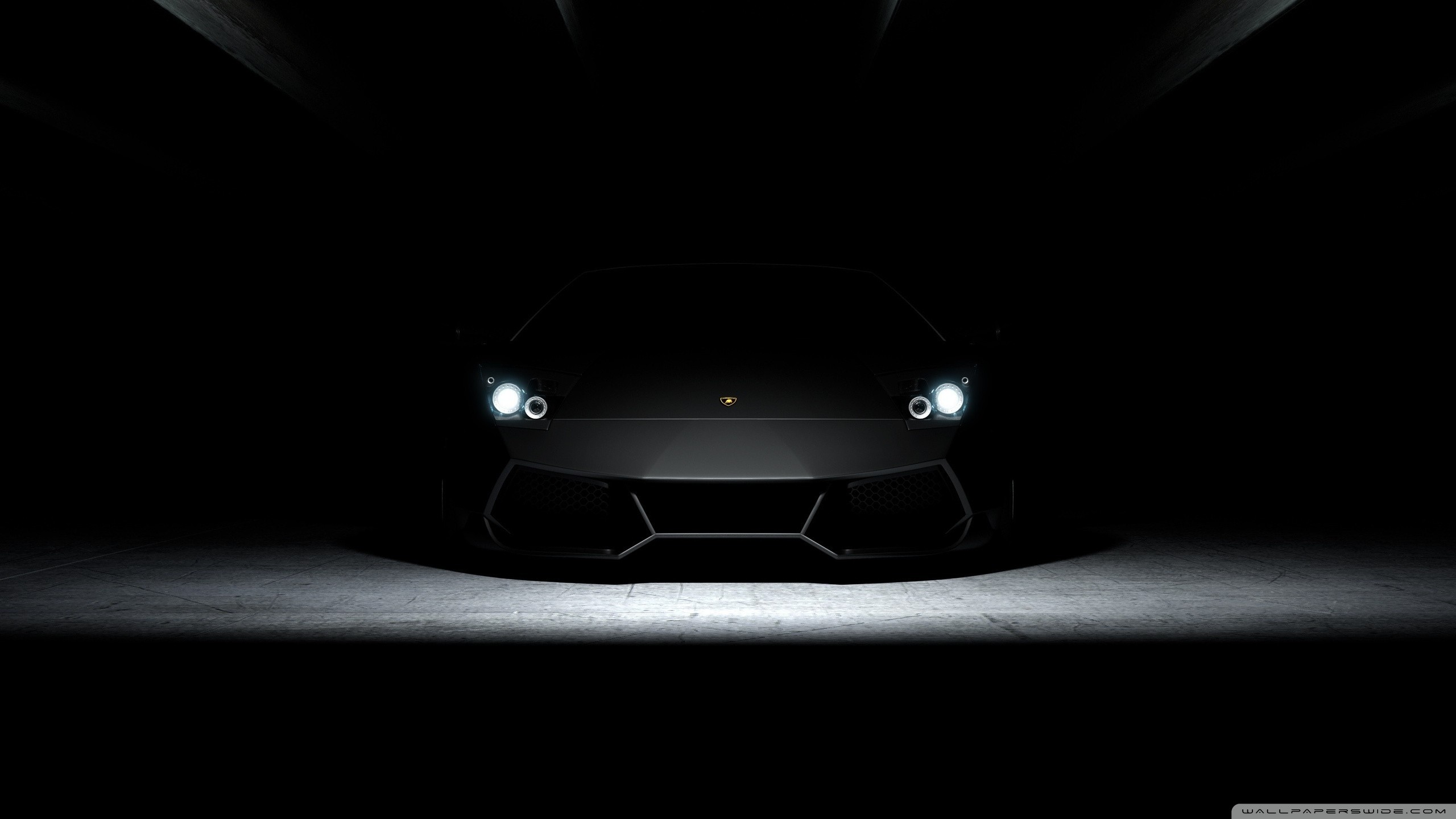 A dark, sleek Lamborghini sports car with its headlights on, parked in a dimly lit garage or parking structure. The car's glossy black finish reflects the minimal light, creating a mysterious and luxurious atmosphere
