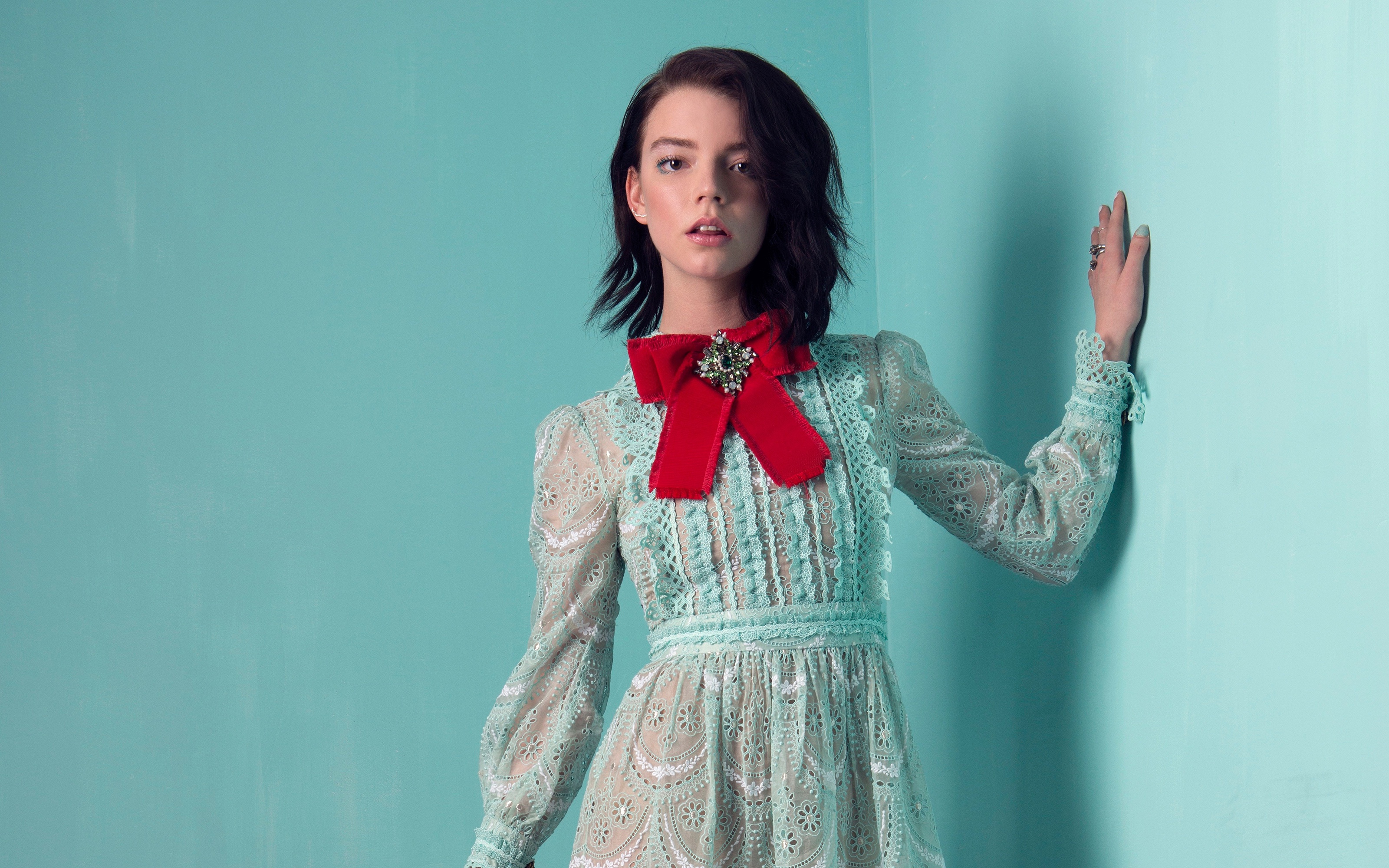 Anya Taylor-Joy posing in a light green lace dress with a red bow against a teal background