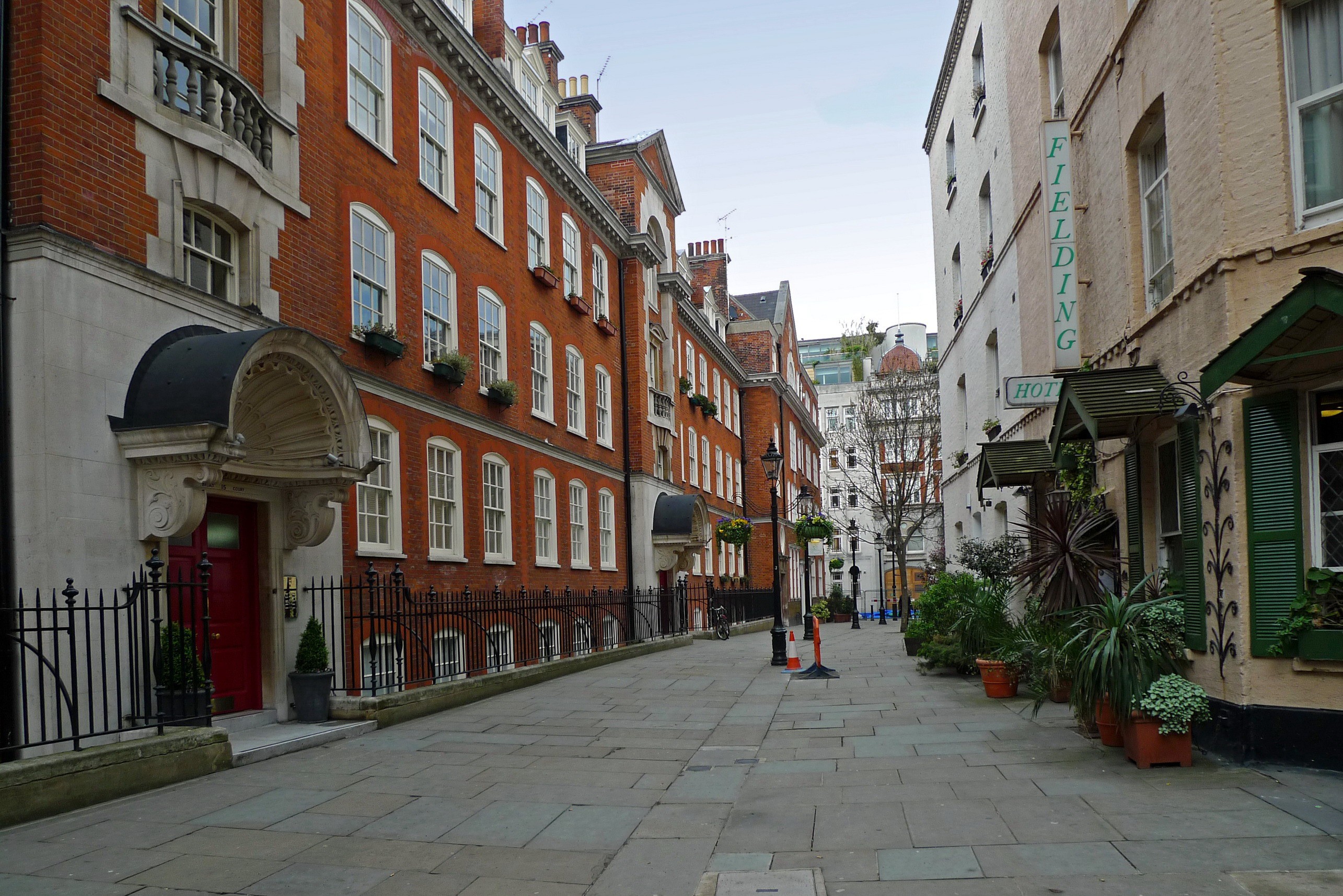 A quiet, narrow street in London with red brick buildings, a cobblestone walkway, and potted plants outside a hotel