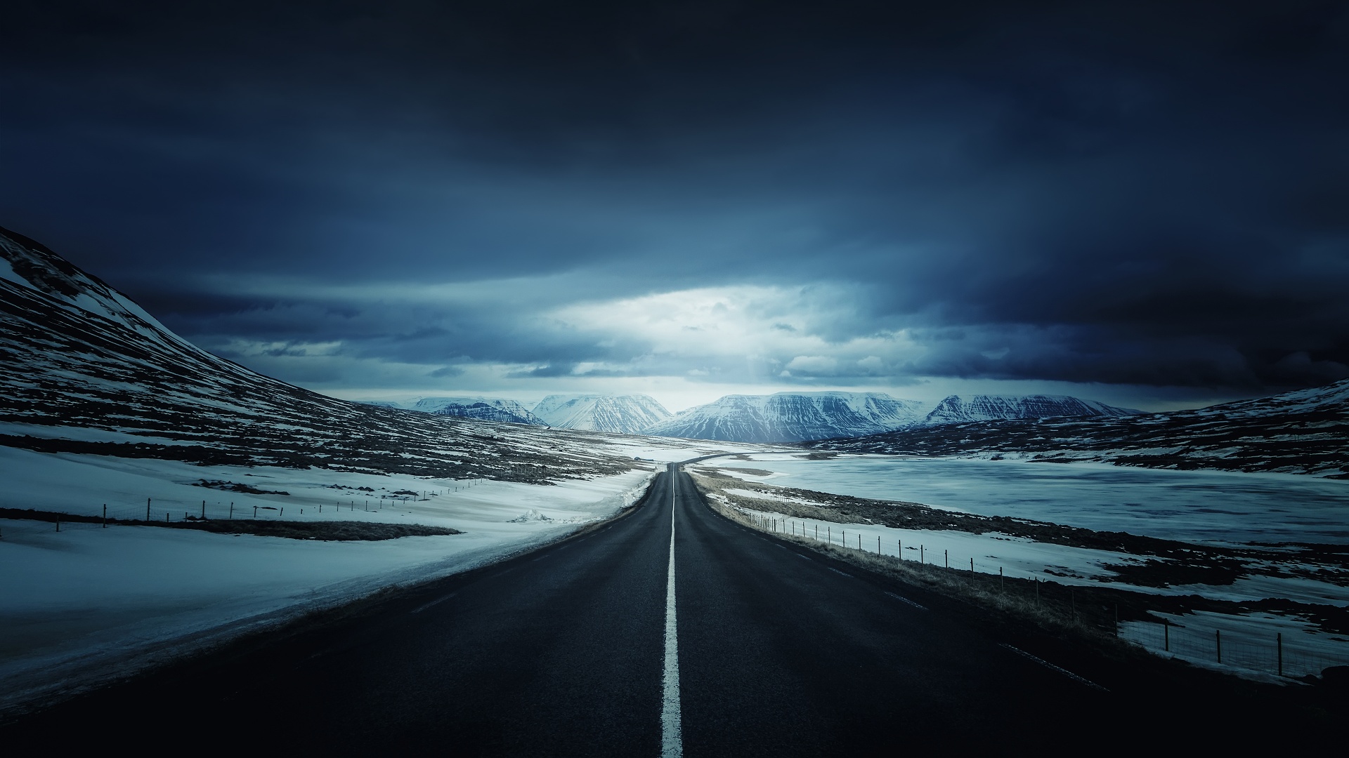 A dark and moody winter landscape with a long, straight road leading towards snow-capped mountains under a dramatic, cloudy sky
