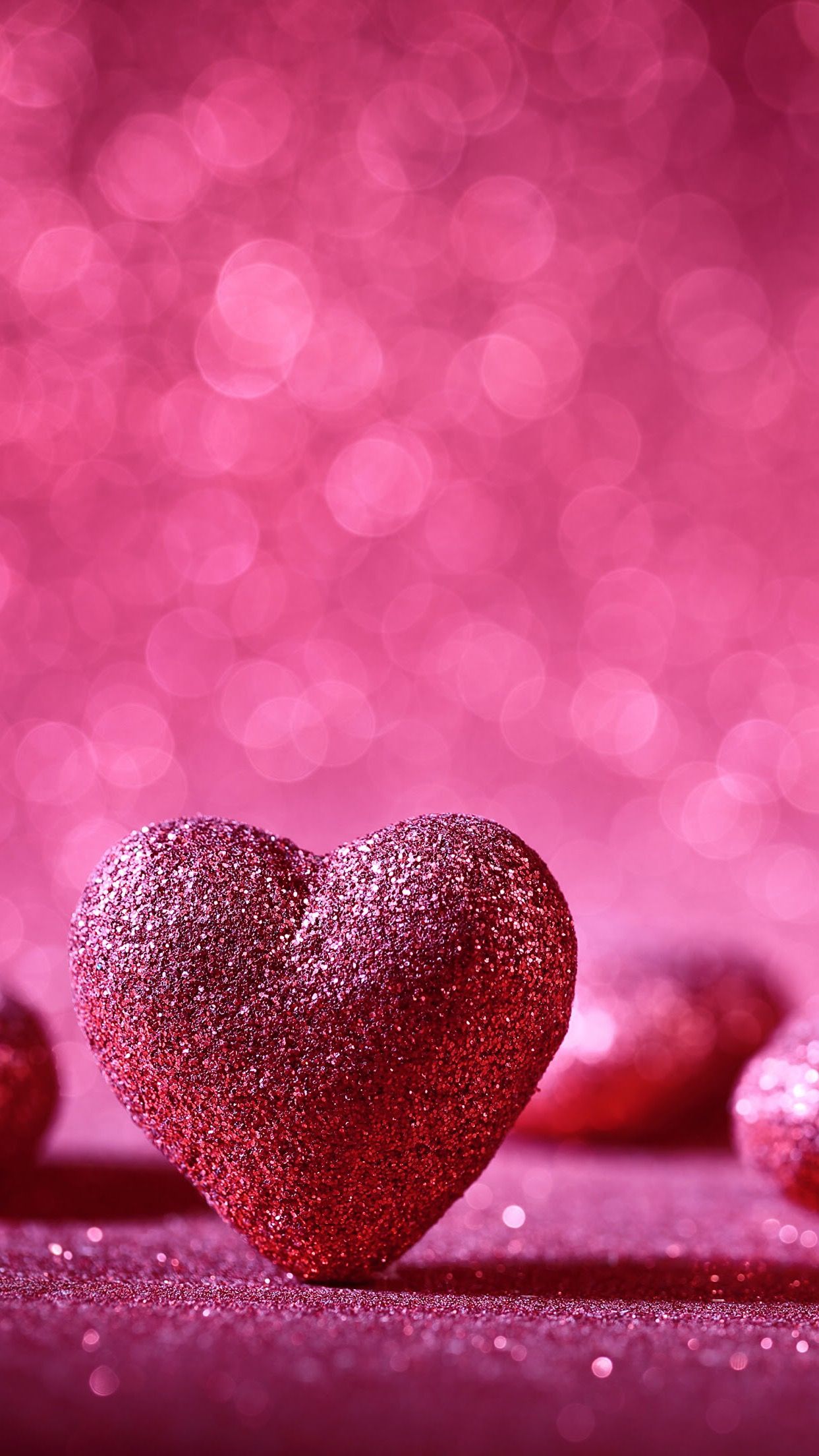 A sparkly red heart sits on a glittery pink surface, with more hearts softly blurred in the background. The backdrop is a bokeh of pink and magenta, creating a romantic and festive atmosphere