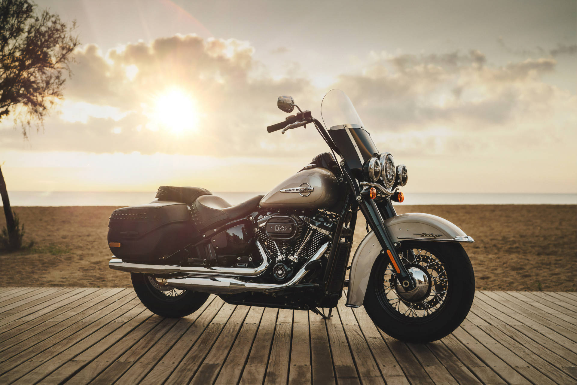 A Harley-Davidson motorcycle parked on a wooden boardwalk at sunset, overlooking a sandy beach and the ocean. The motorcycle is a classic touring model with a windshield and saddlebags, finished in a metallic silver color