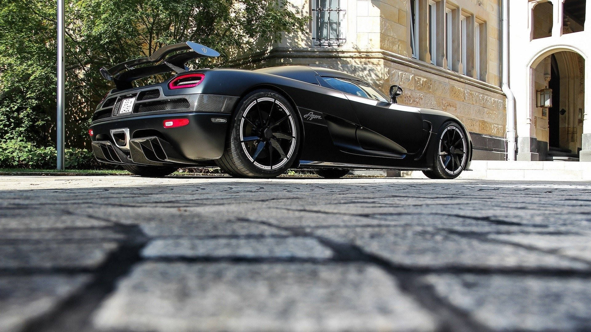 Low-angle view of a matte black Koenigsegg Agera parked on a cobblestone street, highlighting its sleek design and powerful presence