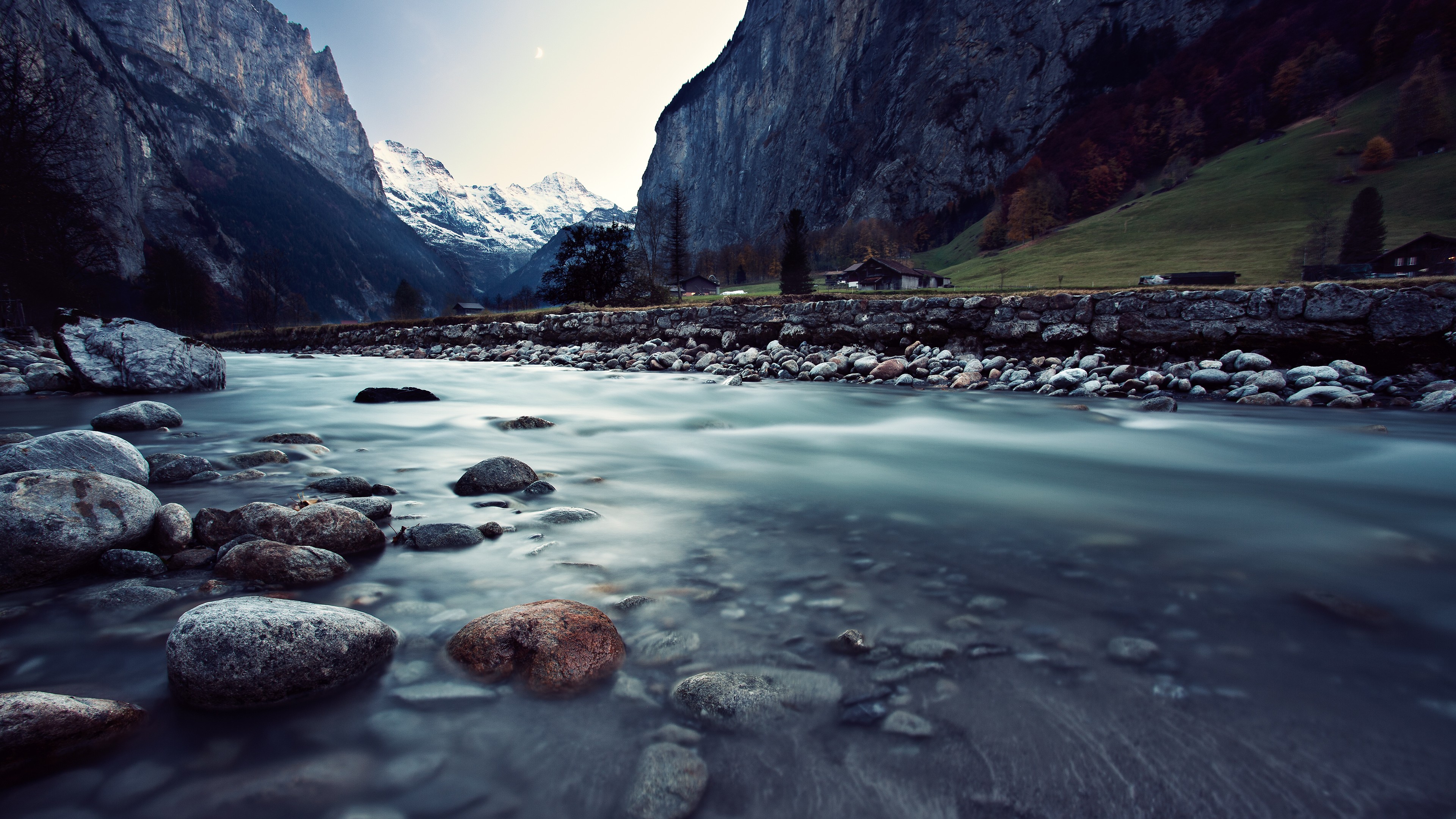 A breathtaking 4K wallpaper showcasing a crystal-clear river flowing through a valley nestled between towering mountains. The smooth, long-exposure effect on the water creates a sense of tranquility, while the snow-capped peaks and lush greenery add to the picturesque scenery