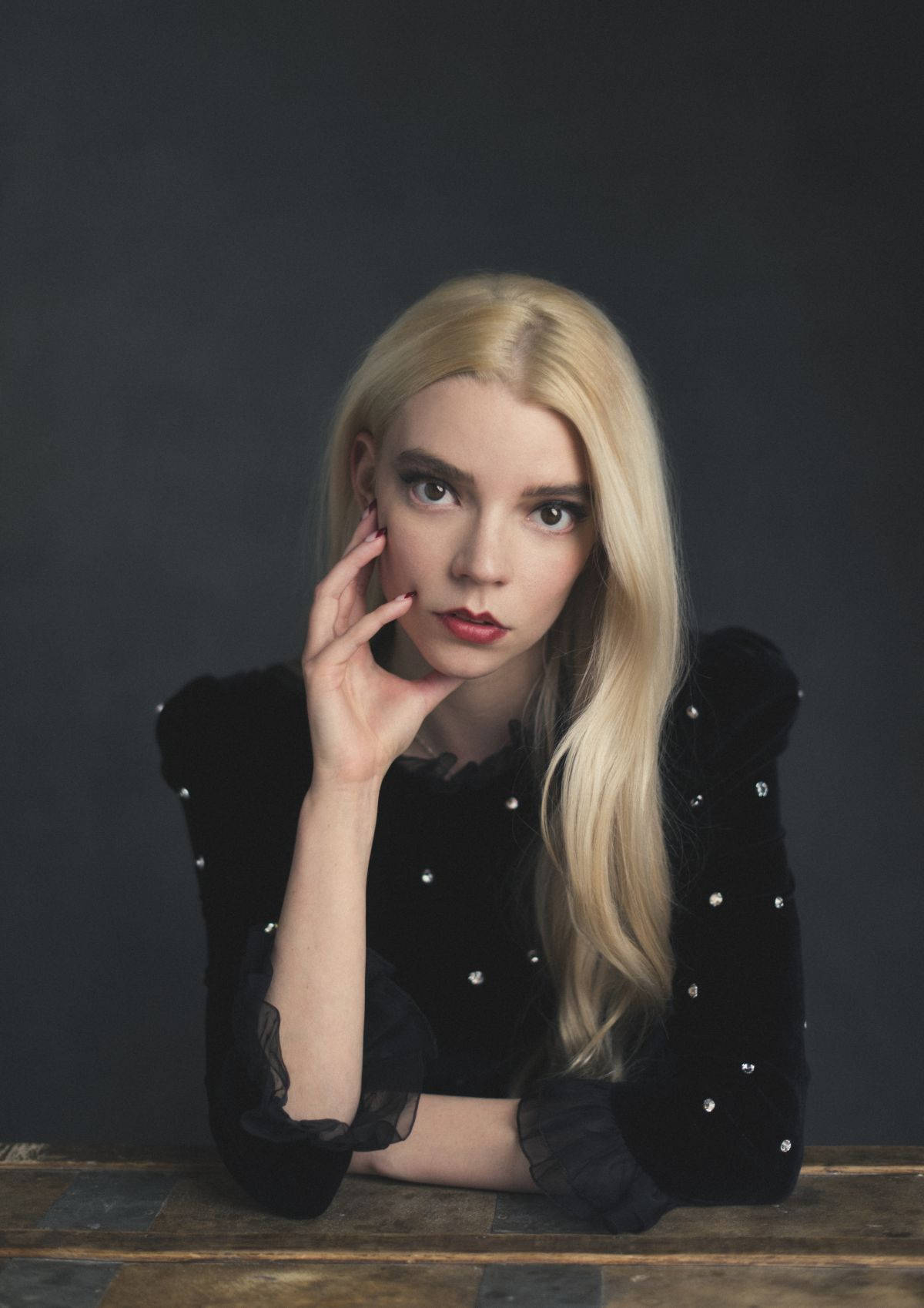 Anya Taylor-Joy posing in a black velvet dress with pearl embellishments, her hand gently touching her face