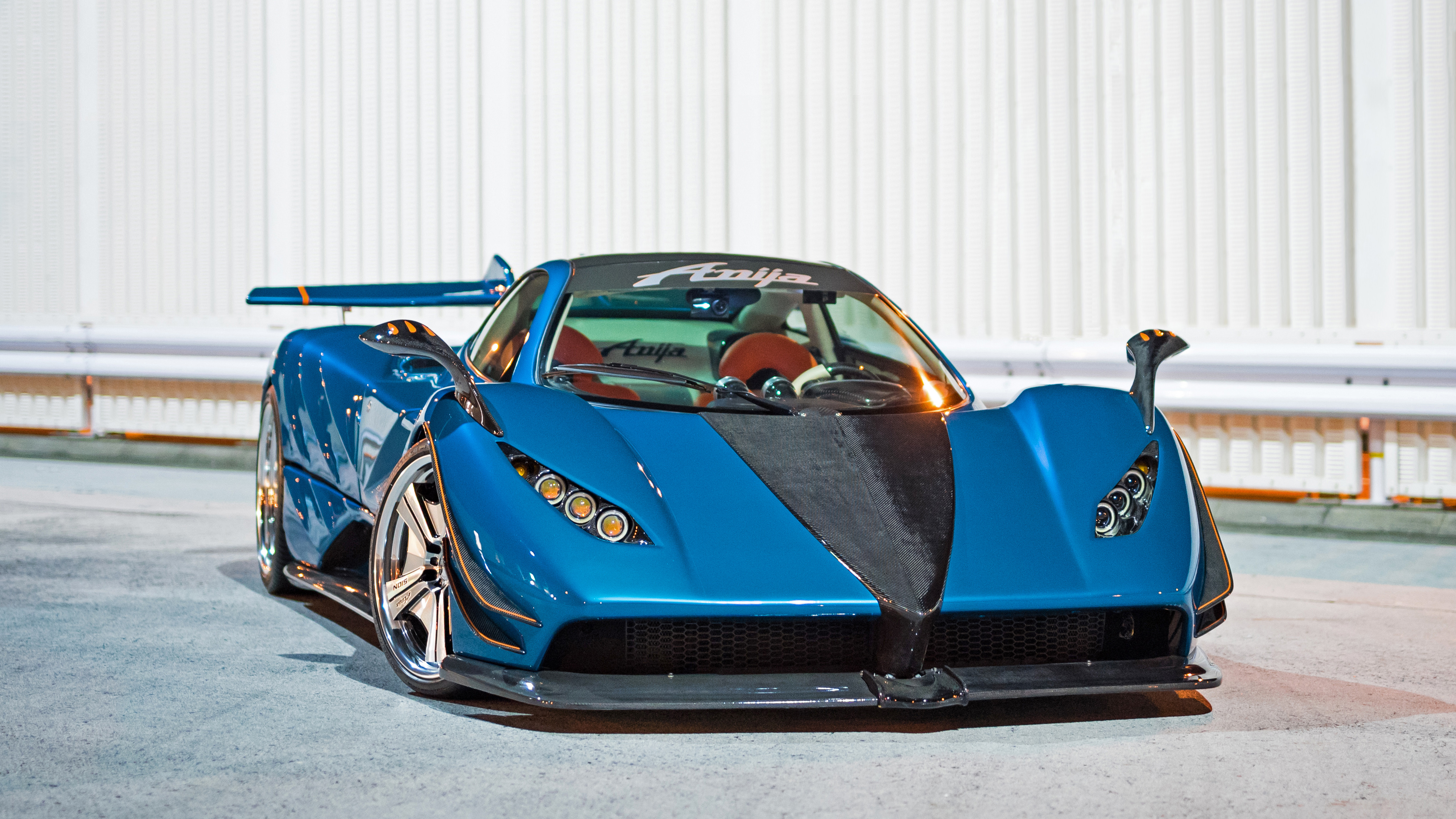 A vibrant blue Pagani Huayra, customized by Anija, parked in front of a white corrugated metal wall. The car's sleek design and carbon fiber accents are highlighted, showcasing its unique and luxurious features