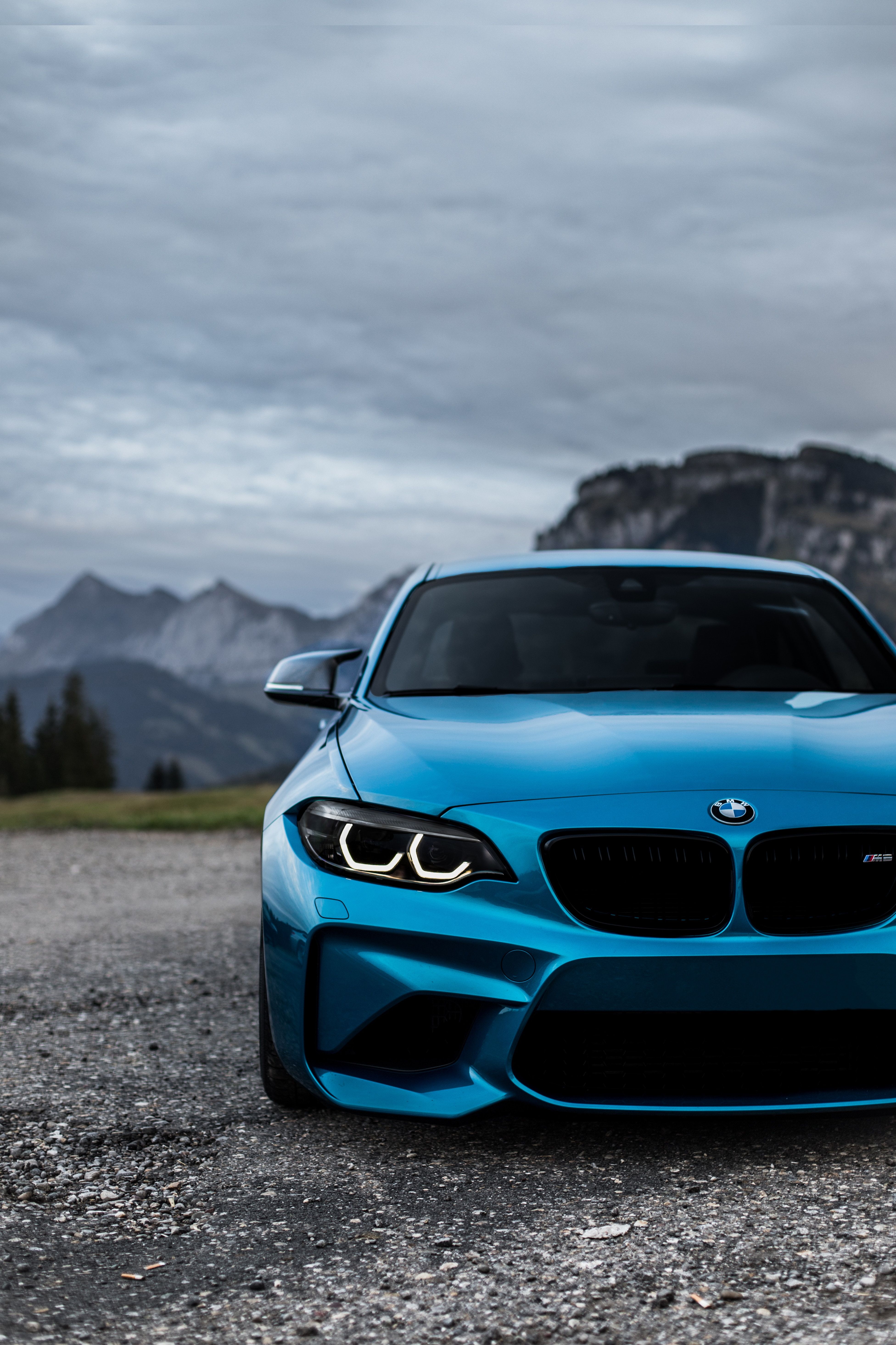 A vibrant blue BMW M2 parked on a gravel road, with a stunning backdrop of majestic mountains and a cloudy sky
