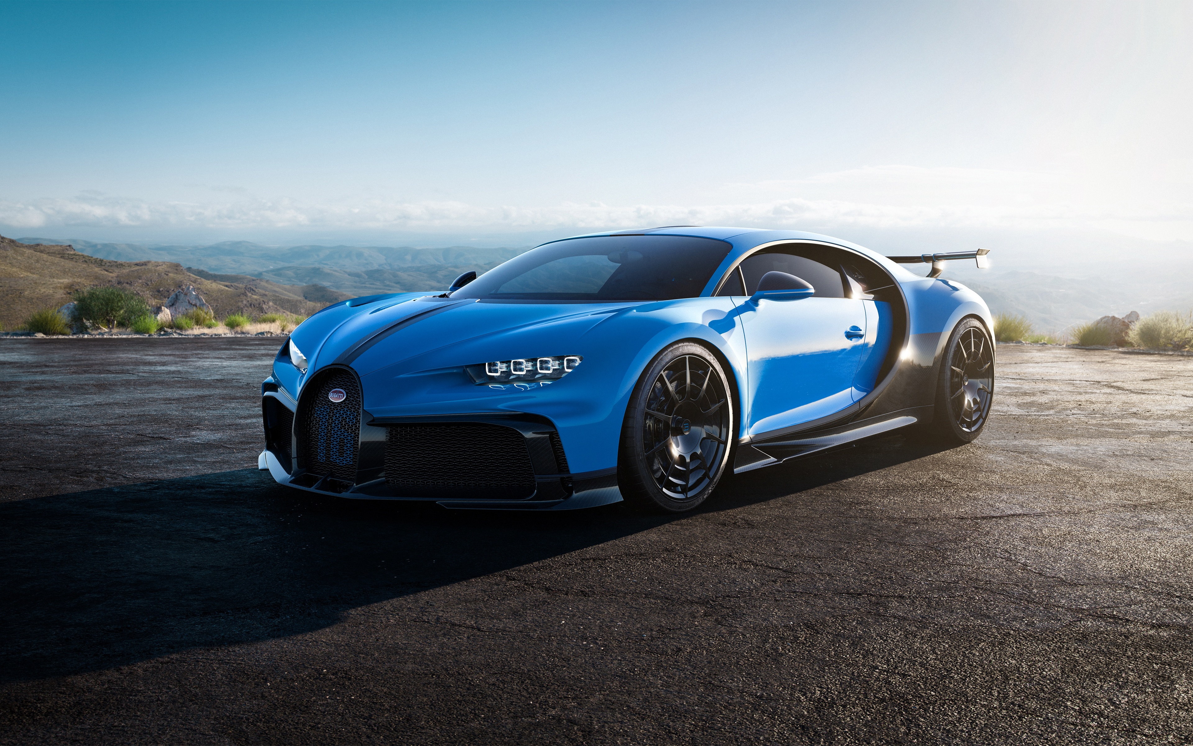 A vibrant blue Bugatti Chiron Super Sport, parked on a mountaintop road, gleams against a backdrop of a picturesque valley and clear sky