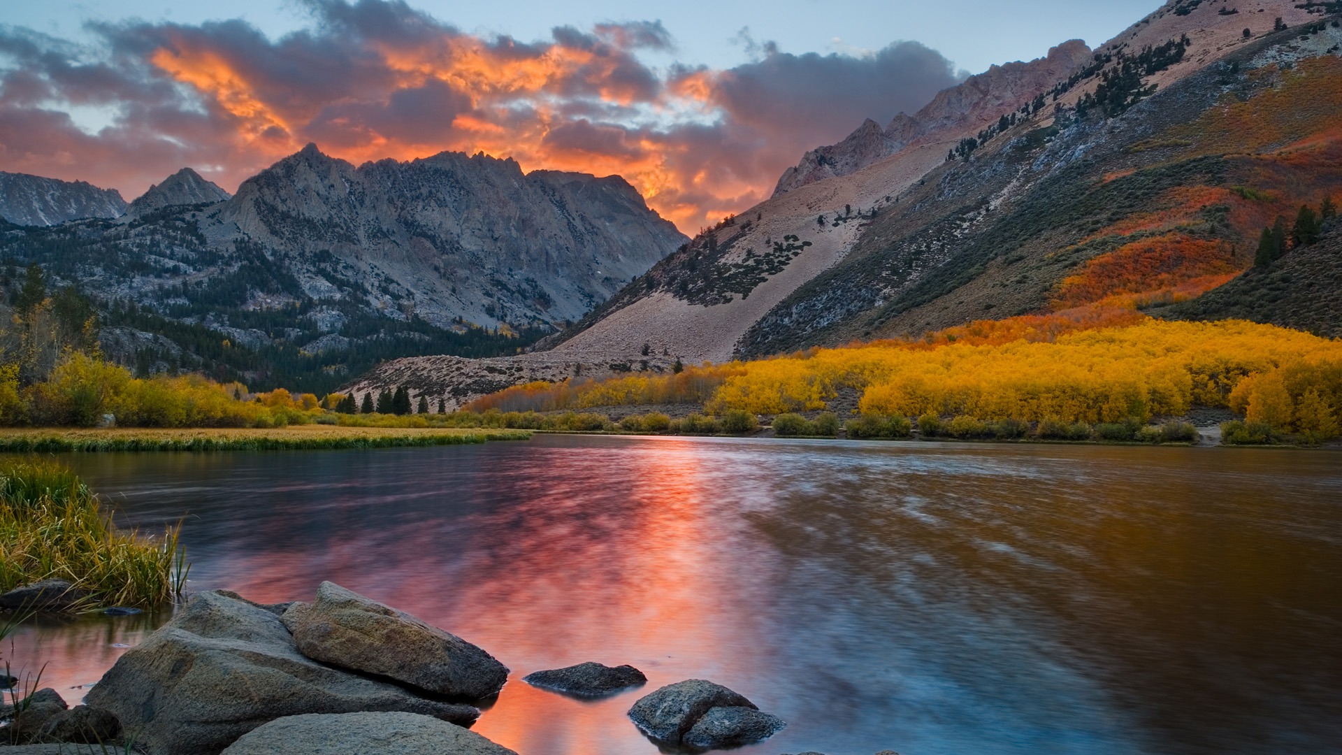 Stunning autumn scenery with colorful trees reflecting in a calm lake, set against a backdrop of majestic mountains and a vibrant sunset sky