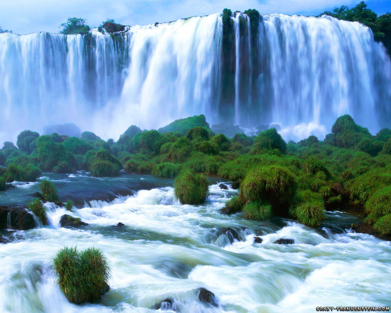 A breathtaking waterfall cascades over a cliff, surrounded by lush green vegetation. The water flows into a turbulent river below, creating a stunning natural landscape