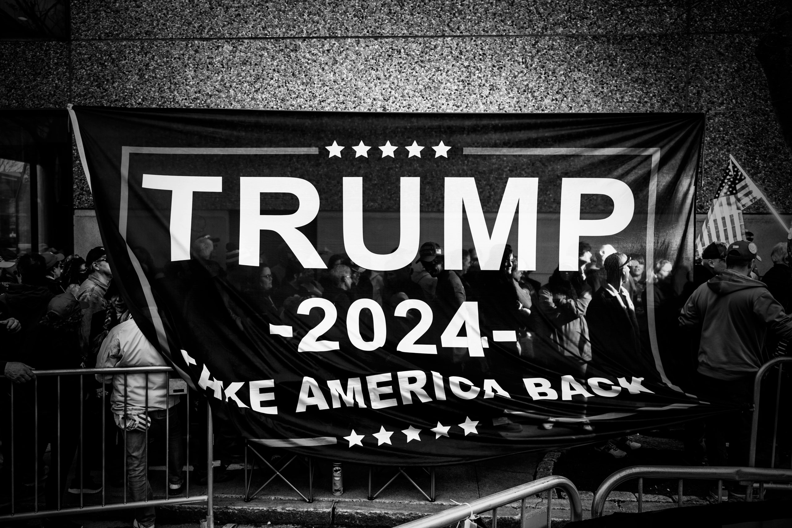 A black and white photo of a crowd of people holding a large banner that reads 'Trump 2024 Take America Back'