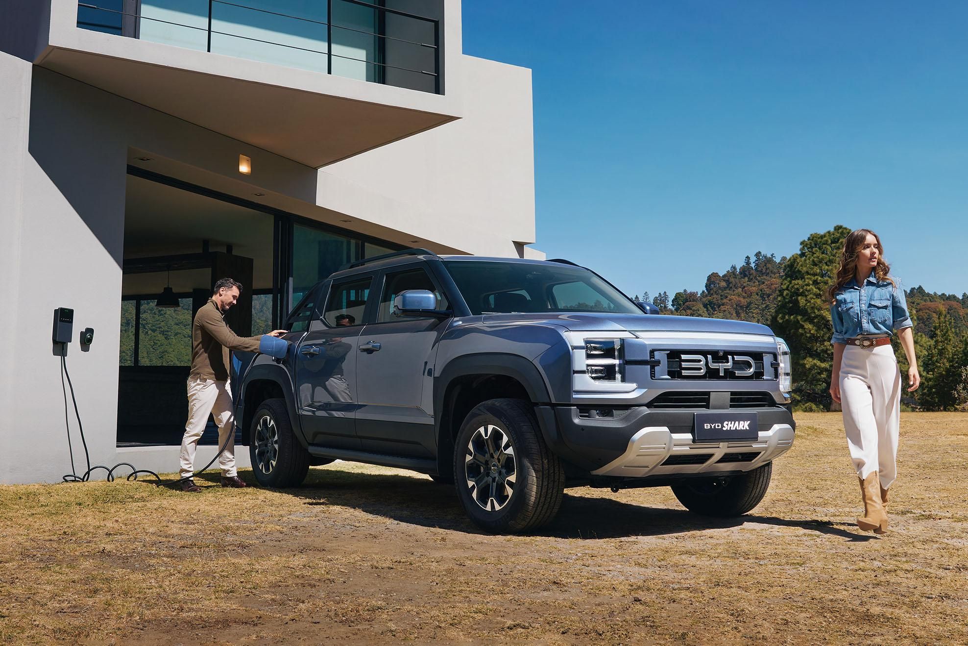 A silver BYD Shark electric pickup truck parked in front of a modern house, being charged by a man while a woman walks nearby