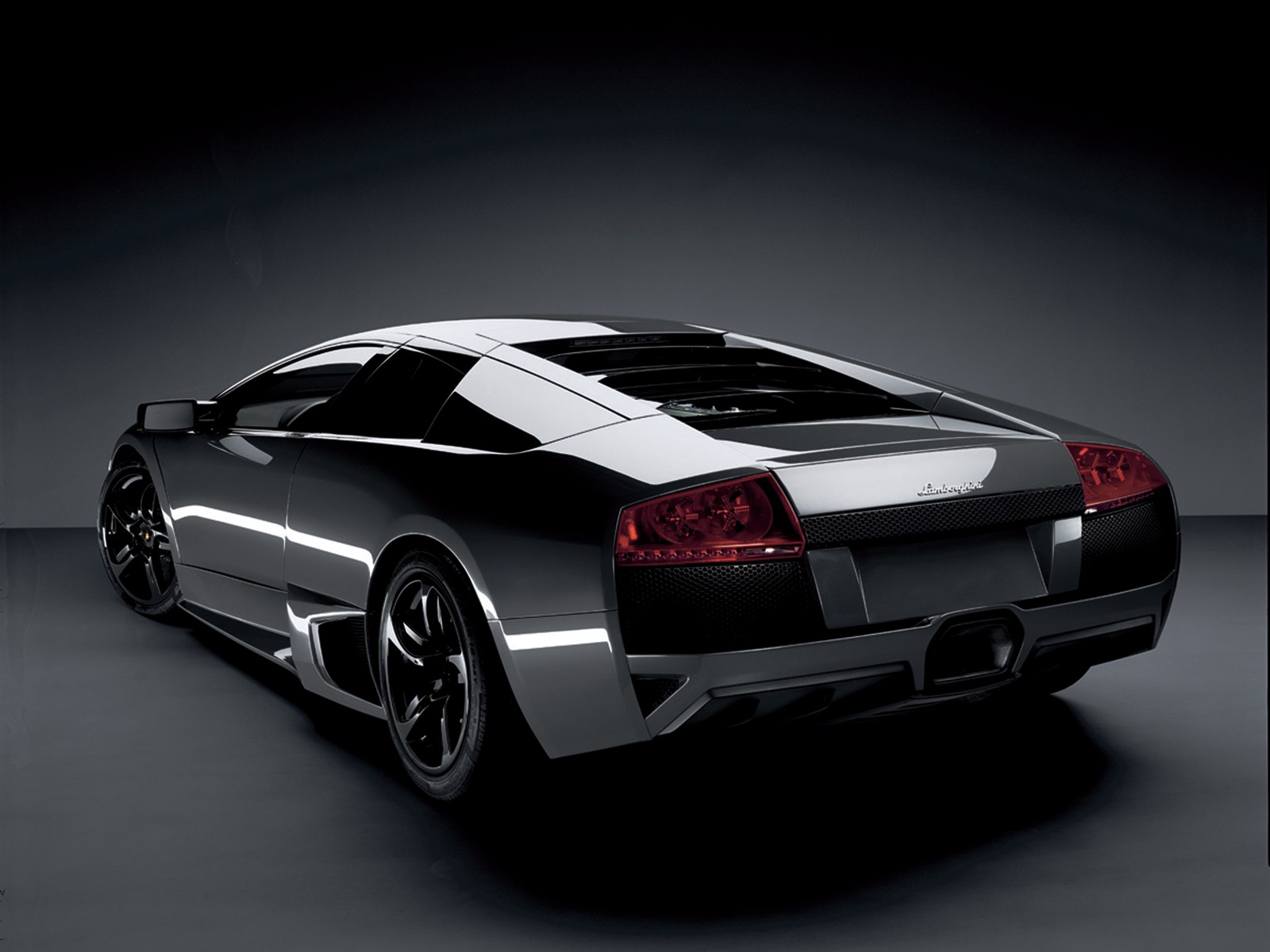 A sleek, silver Lamborghini Murciélago, viewed from the rear, parked against a dark backdrop. Its taillights glow, highlighting the car's powerful design