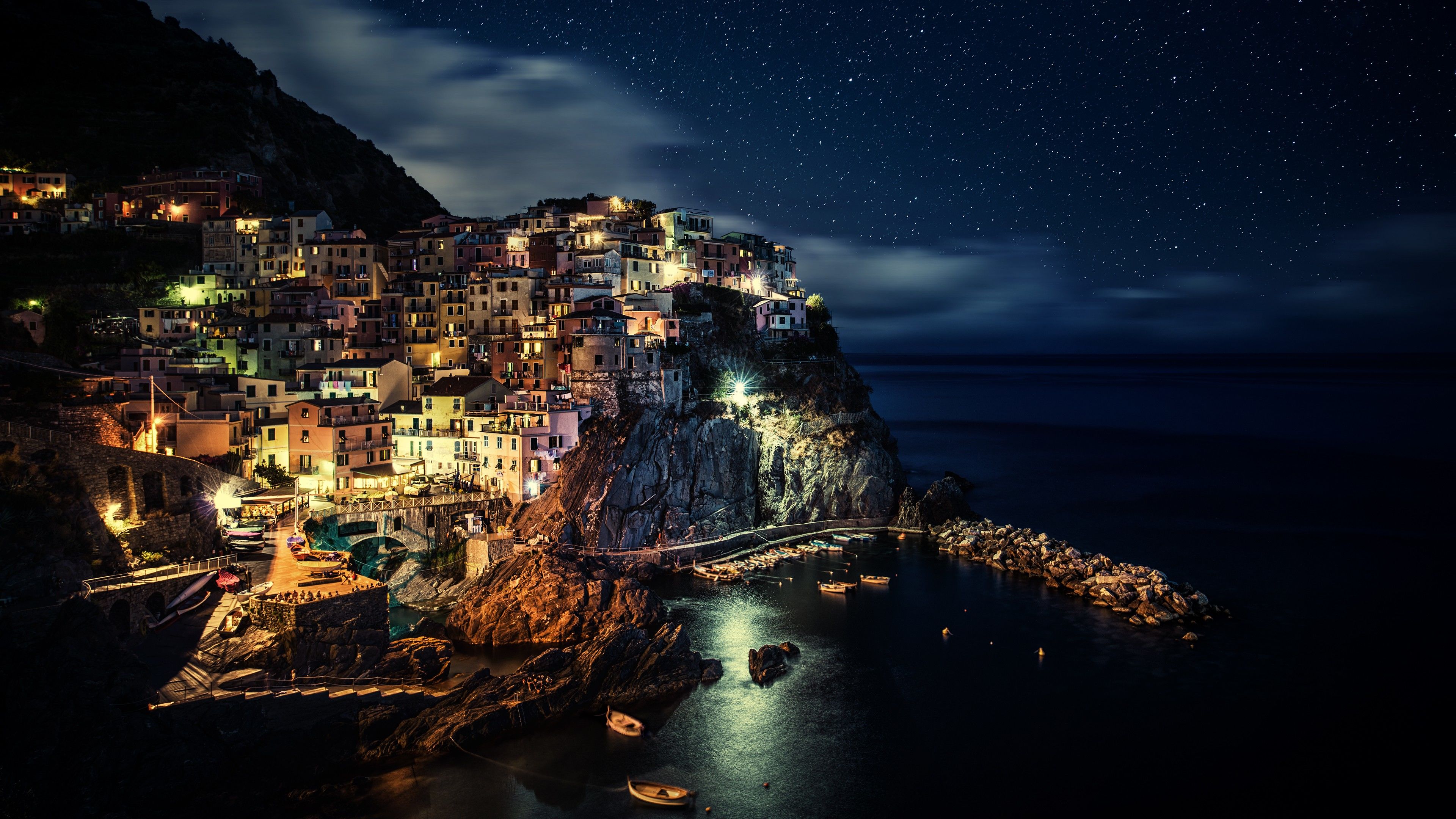 A breathtaking 4K wallpaper of a picturesque Italian village perched on a cliff overlooking the sea at night, illuminated by warm lights under a starry sky