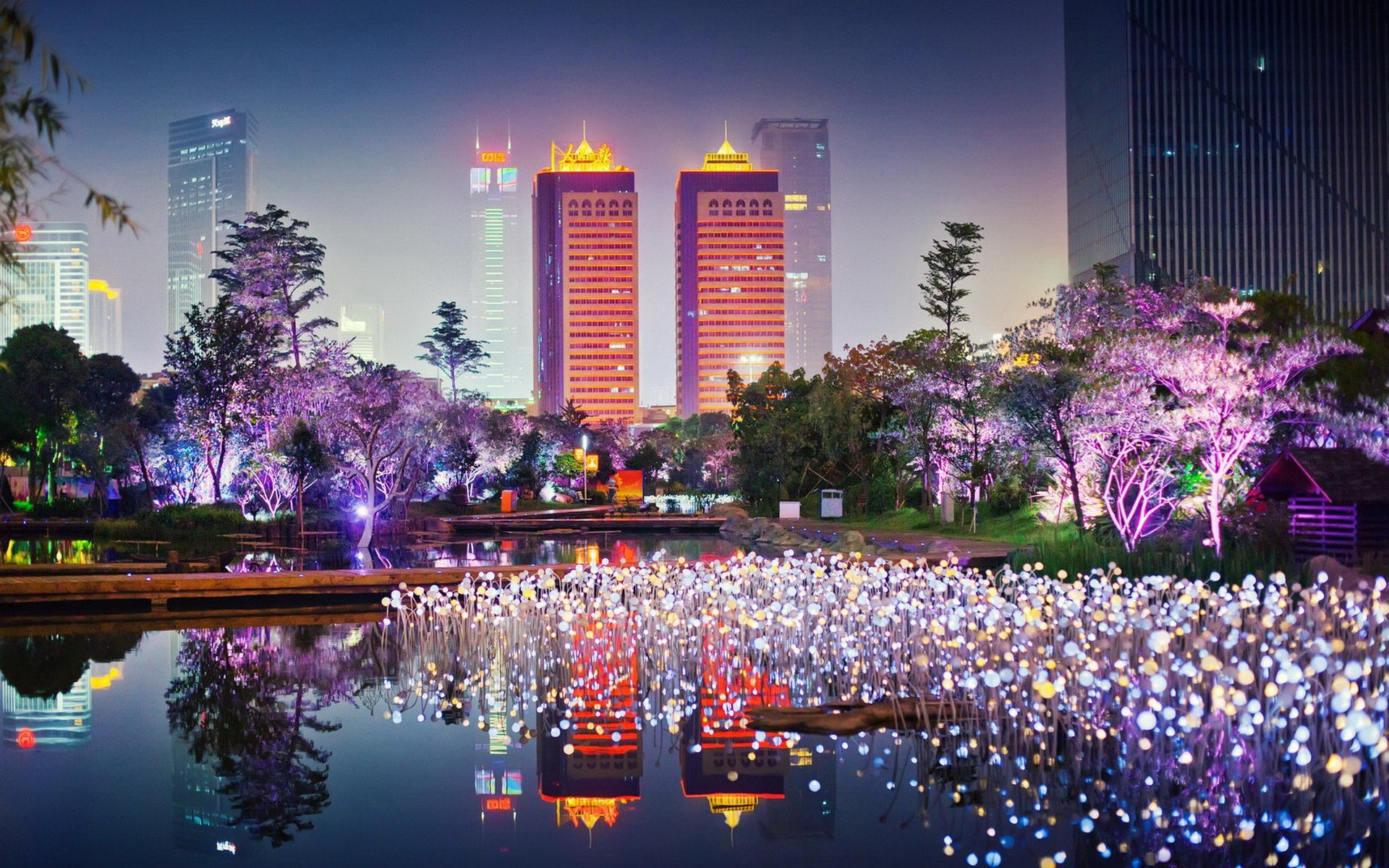 Vibrant cityscape at night with illuminated buildings and a light display in a park reflecting on a pond