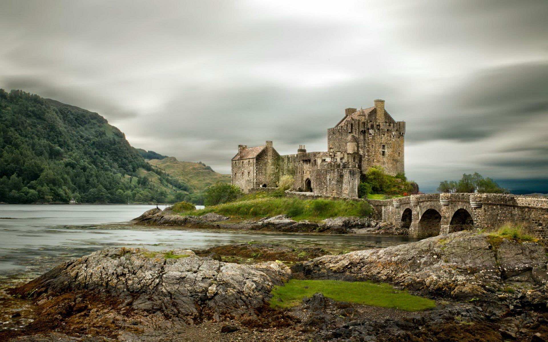 A majestic medieval castle stands on a small island, connected to the mainland by a stone bridge. Lush green vegetation covers the island, contrasting with the castle's gray stone walls. The surrounding landscape features rocky shores, calm waters, and a backdrop of rolling hills under a dramatic, cloudy sky