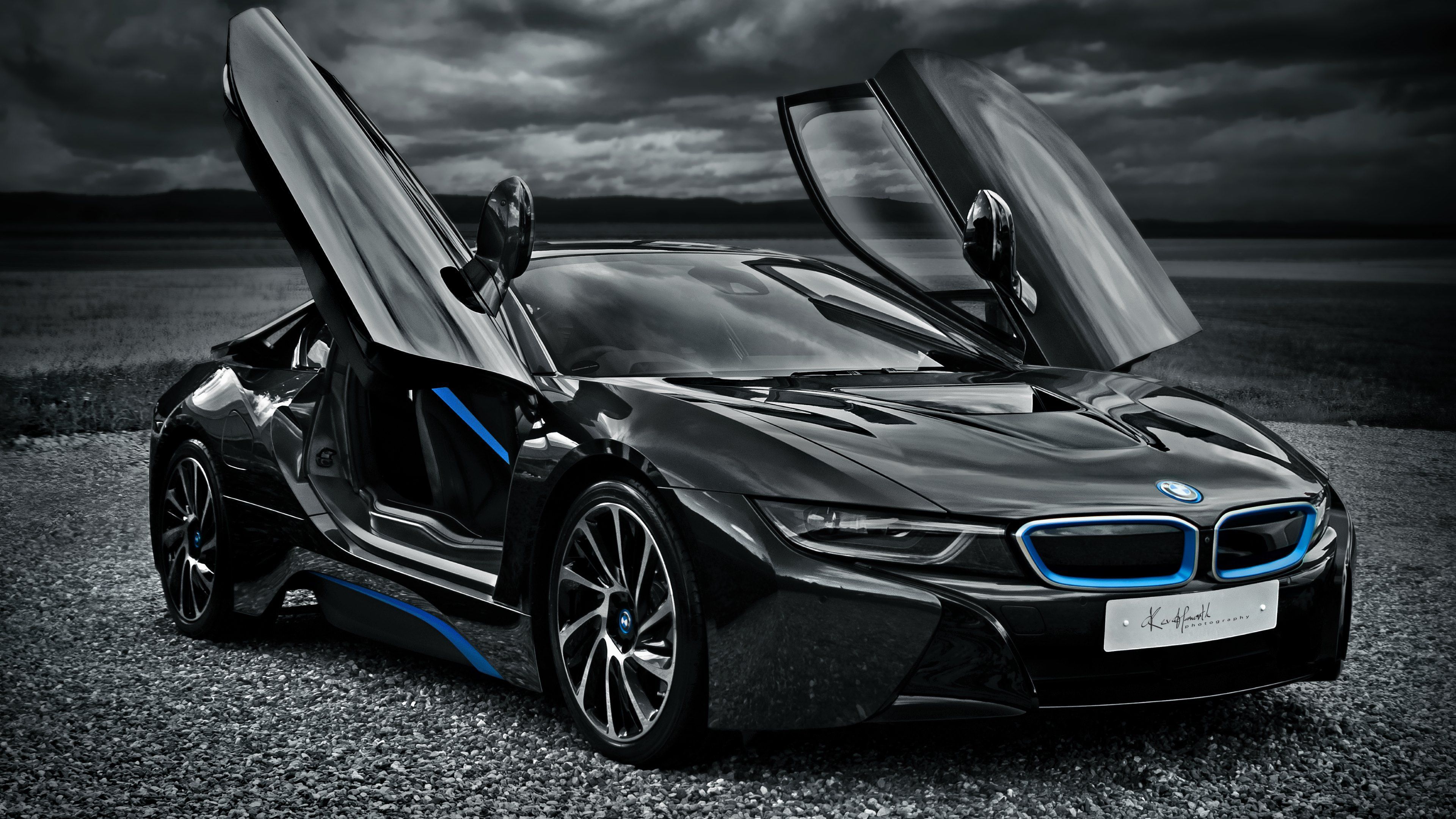 A sleek black BMW i8 sports car with scissor doors open, parked on a gravel road against a dramatic sky backdrop