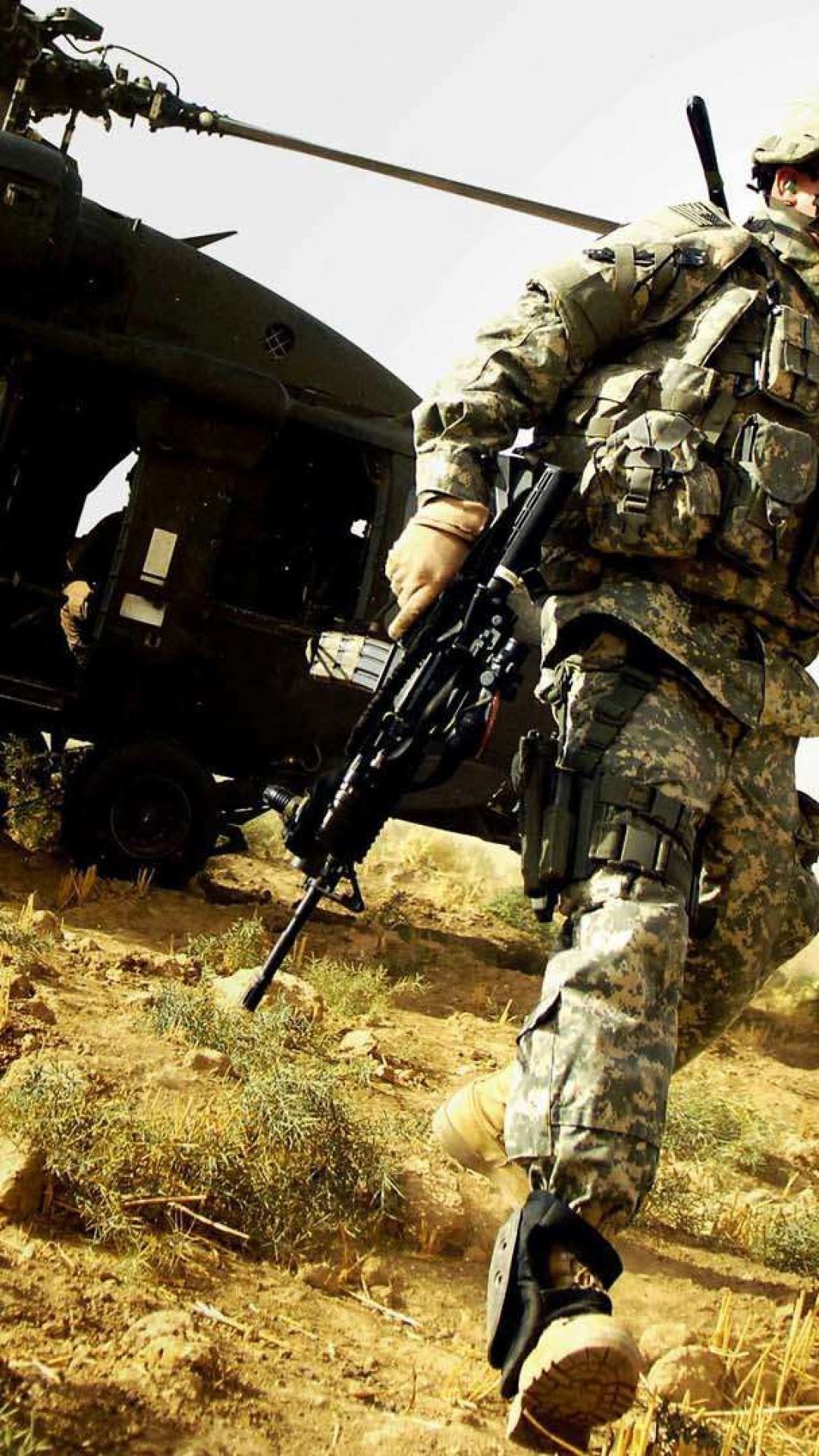 A soldier in camouflage uniform disembarks from a military helicopter in a desert environment, carrying a rifle