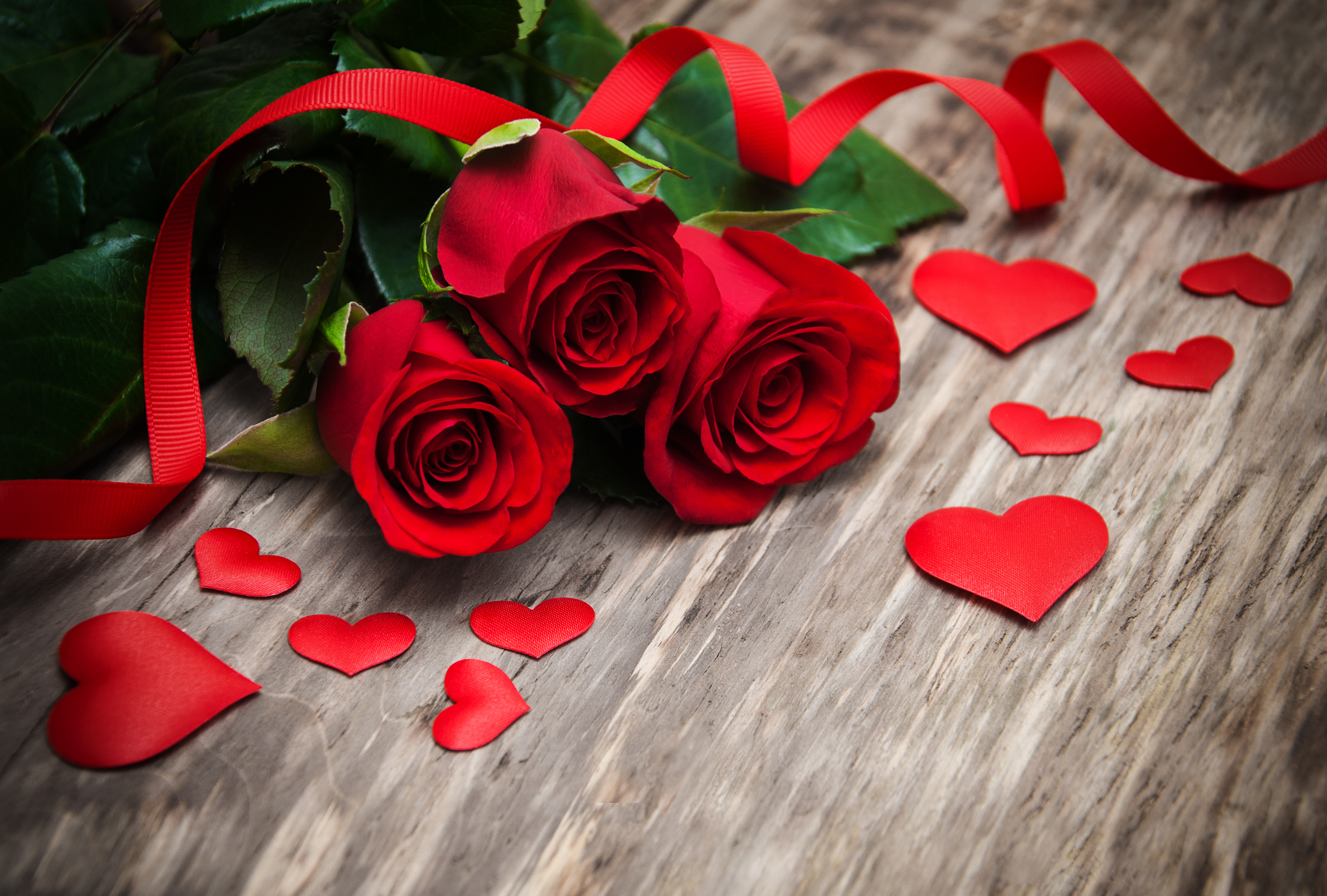 Three red roses with a red ribbon and heart confetti on a wooden table, perfect for Valentine's Day