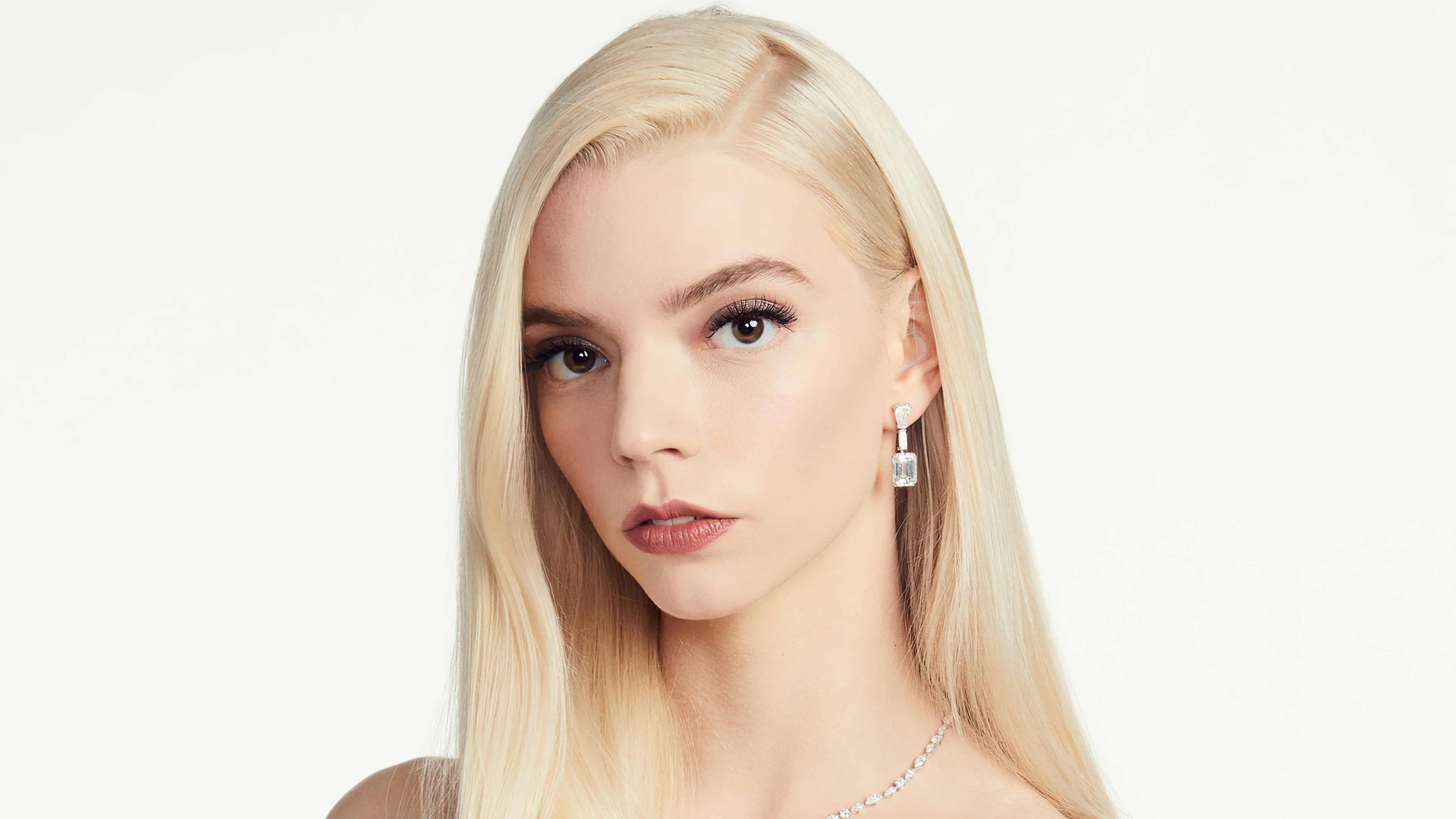 Close-up portrait of Anya Taylor-Joy with blonde hair, styled straight and cascading over her shoulders, and wearing elegant diamond earrings. She gazes directly at the camera against a clean white background