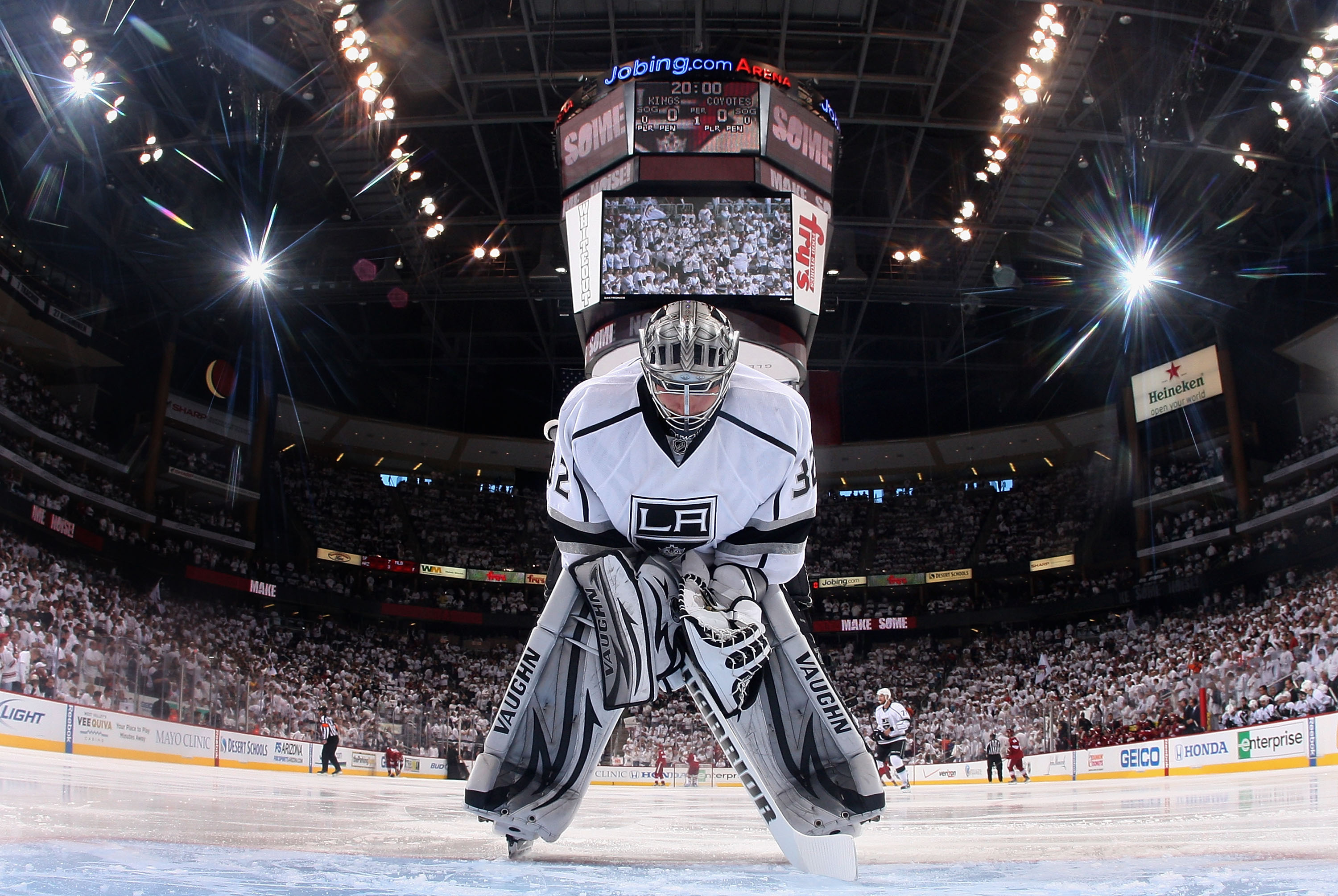 A hockey goalie in full gear crouches on the ice, focused on the game, in a brightly lit arena with a large scoreboard overhead