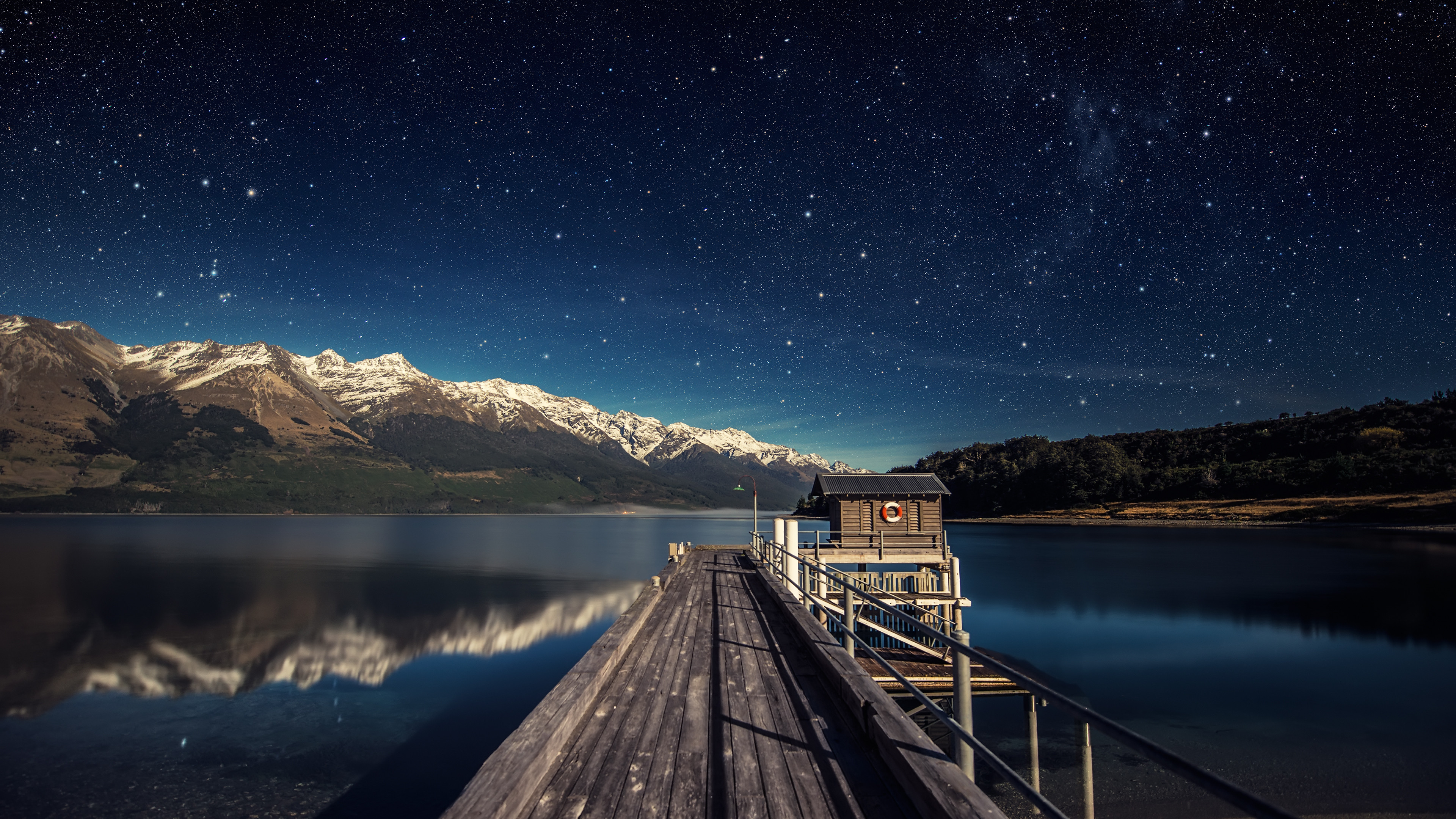 A serene 4K HD wallpaper featuring a wooden pier extending into a tranquil lake, with majestic snow-capped mountains reflected in the water under a breathtaking starry night sky