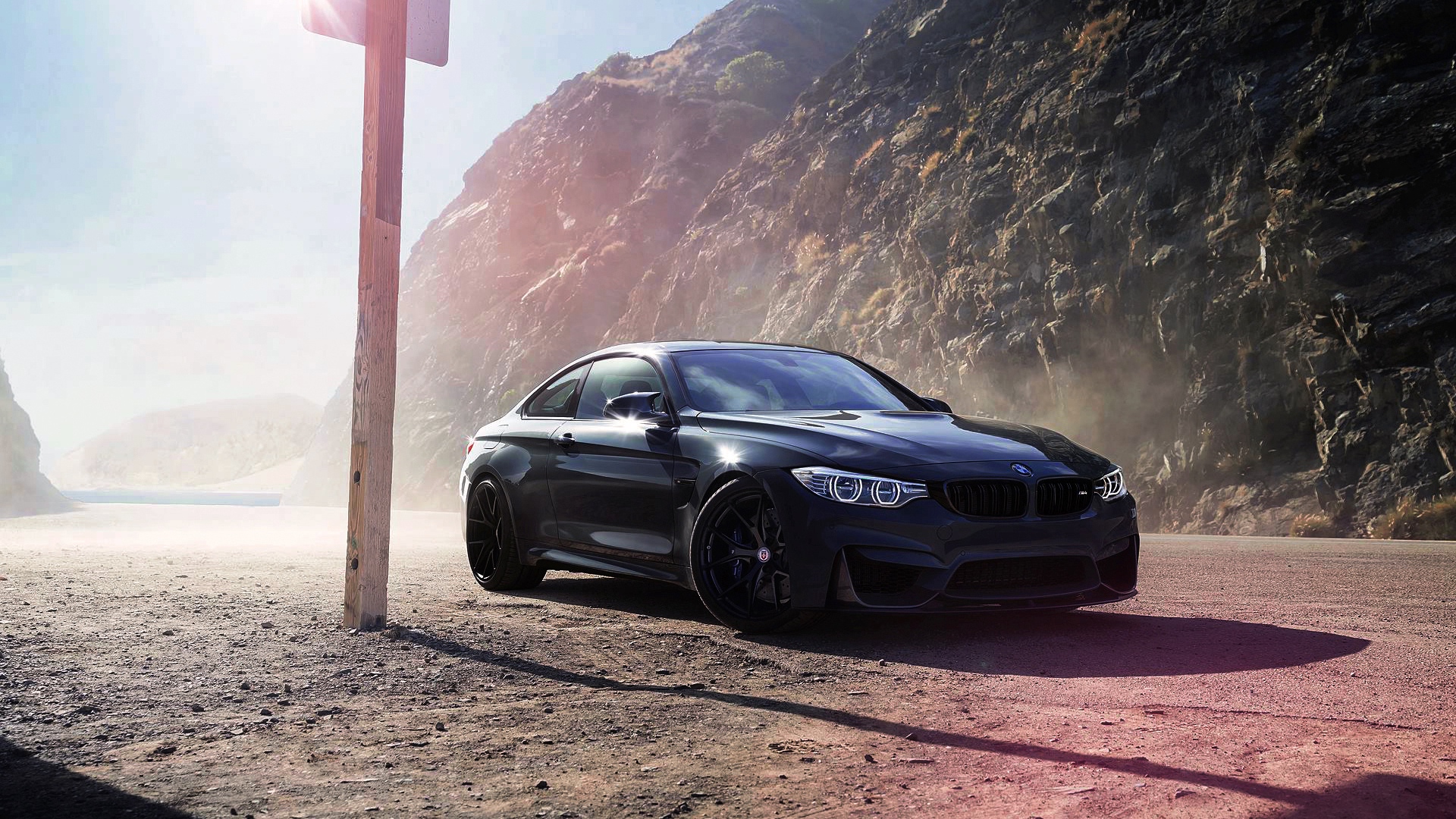 A sleek, black BMW M4 coupe parked on a dusty road with a rocky cliff in the background. The sun is shining brightly, creating a dramatic contrast between the car and the landscape